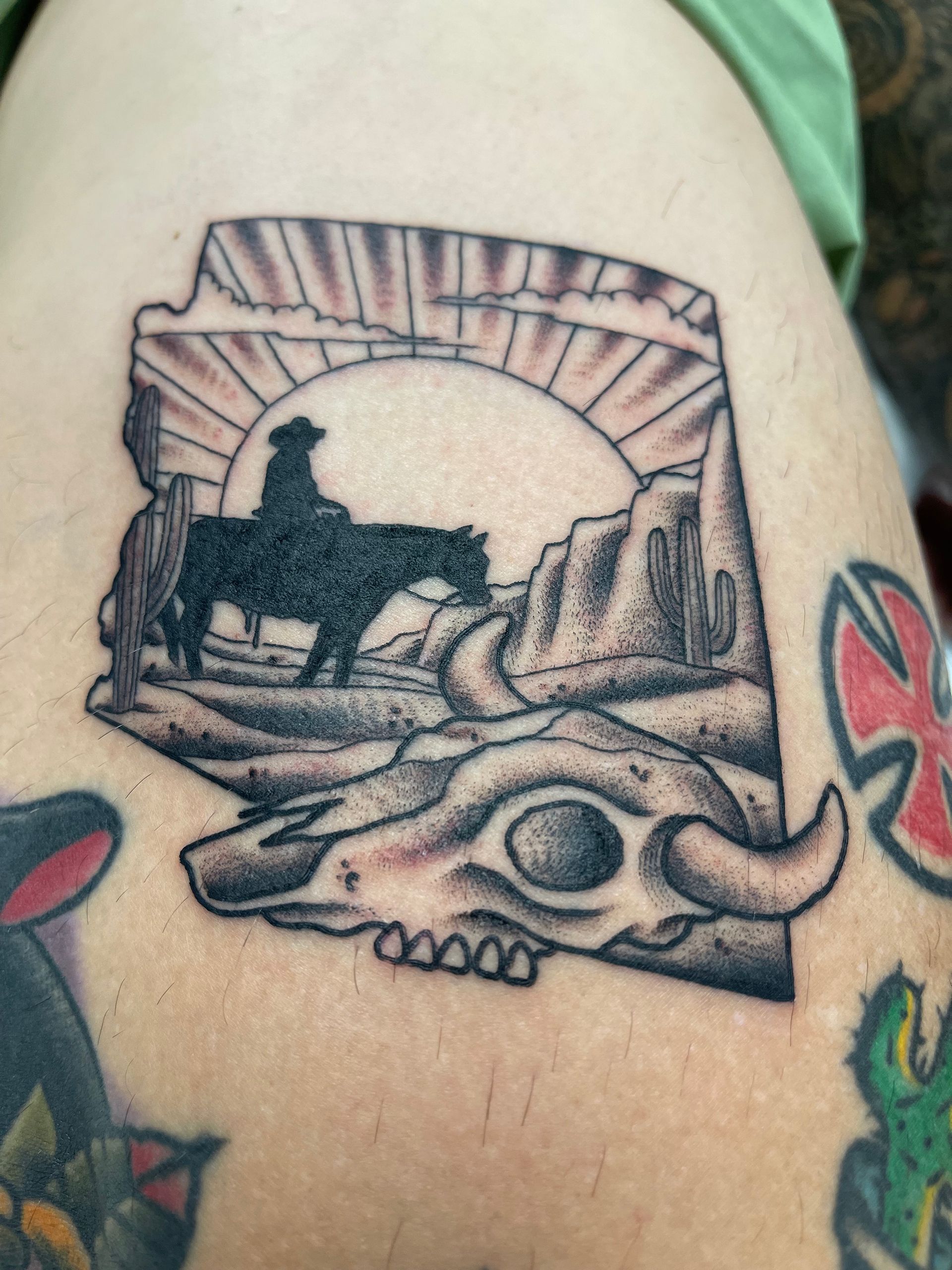 Arizona state outline tattoo with cowboy on horse, skull, desert landscape, and sun.