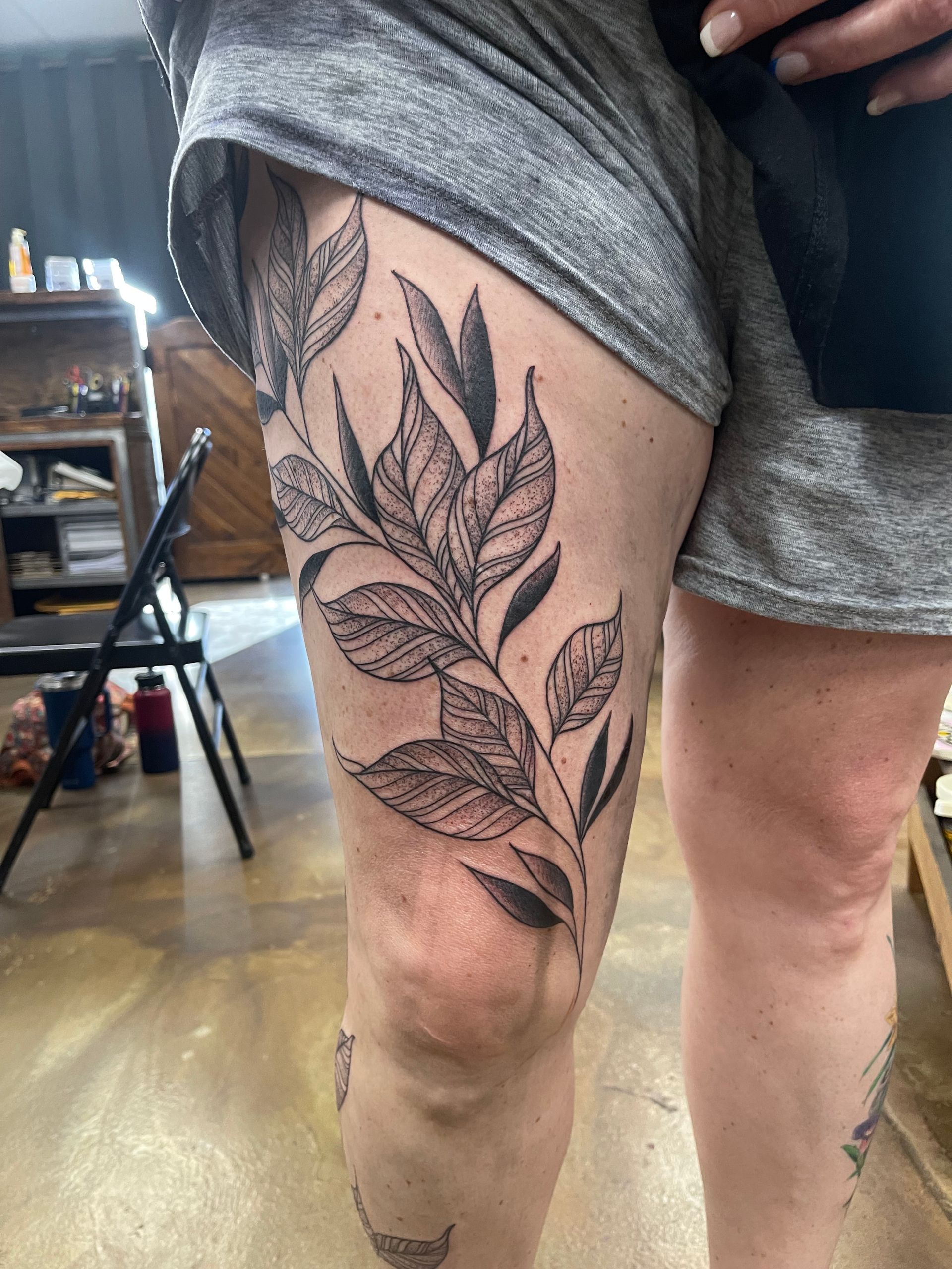 Tattoo of leaves and branches on a person's thigh. Gray leaves, gray shorts. Wooden floor.
