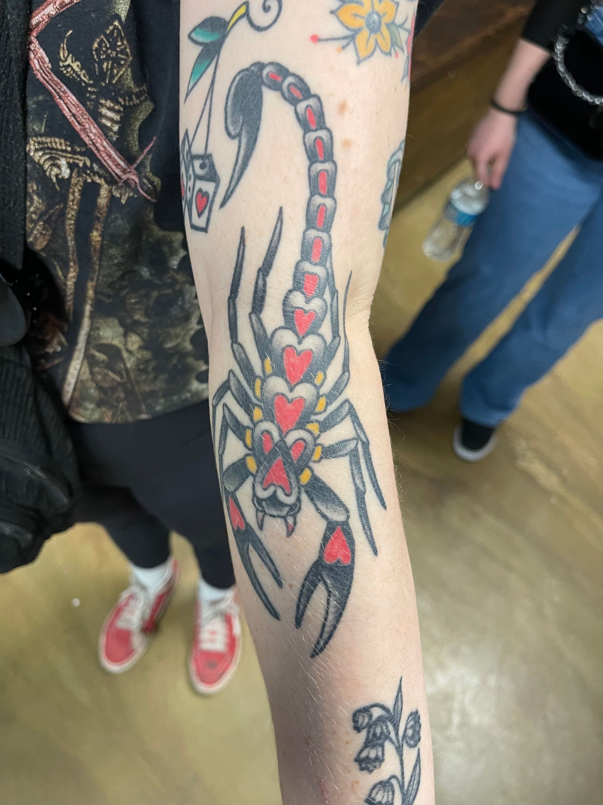 Arm tattoo of a scorpion with a heart-shaped body and red accents. Other tattoos visible.