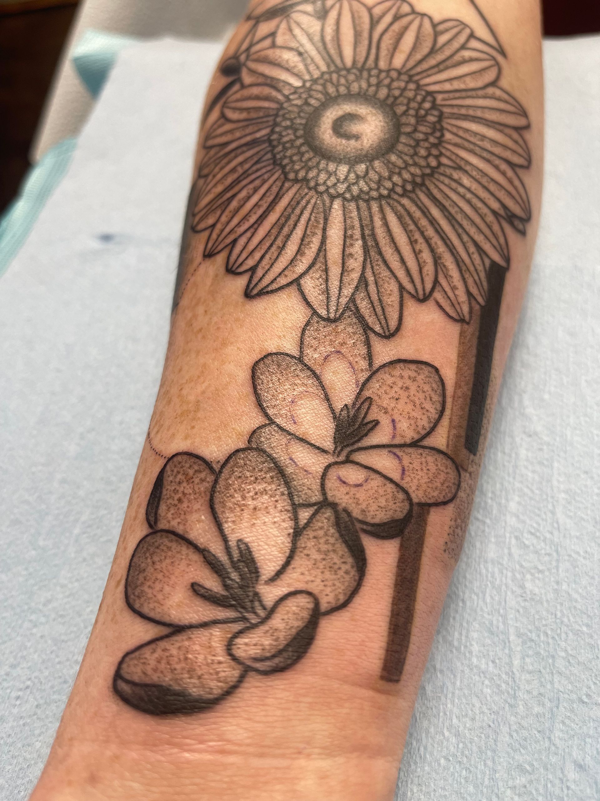 Tattoo on a forearm; a sunflower above two flowers, with a vertical black line.
