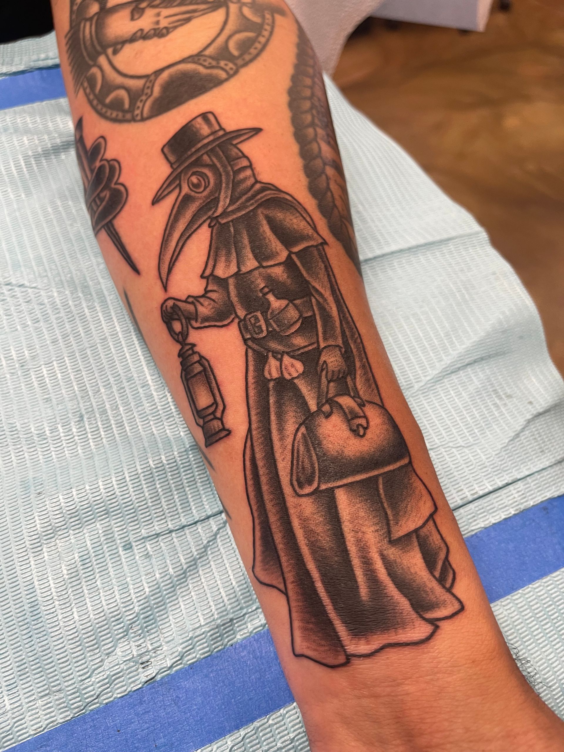 Tattoo of a plague doctor in black ink, holding a lantern and bag on an arm.