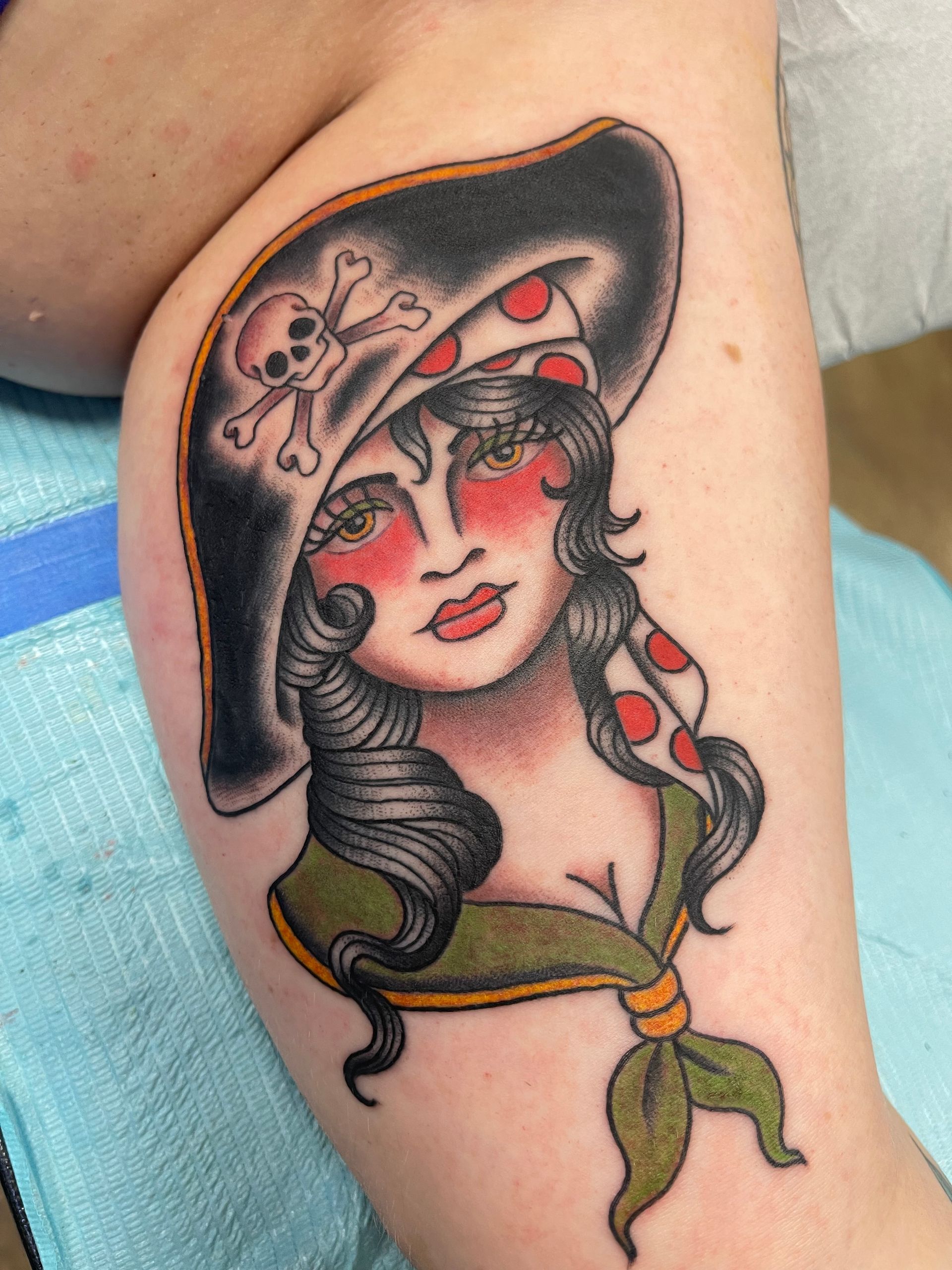 Traditional tattoo of a woman with red cheeks, wearing a pirate hat with skull, bandanna, and green scarf.