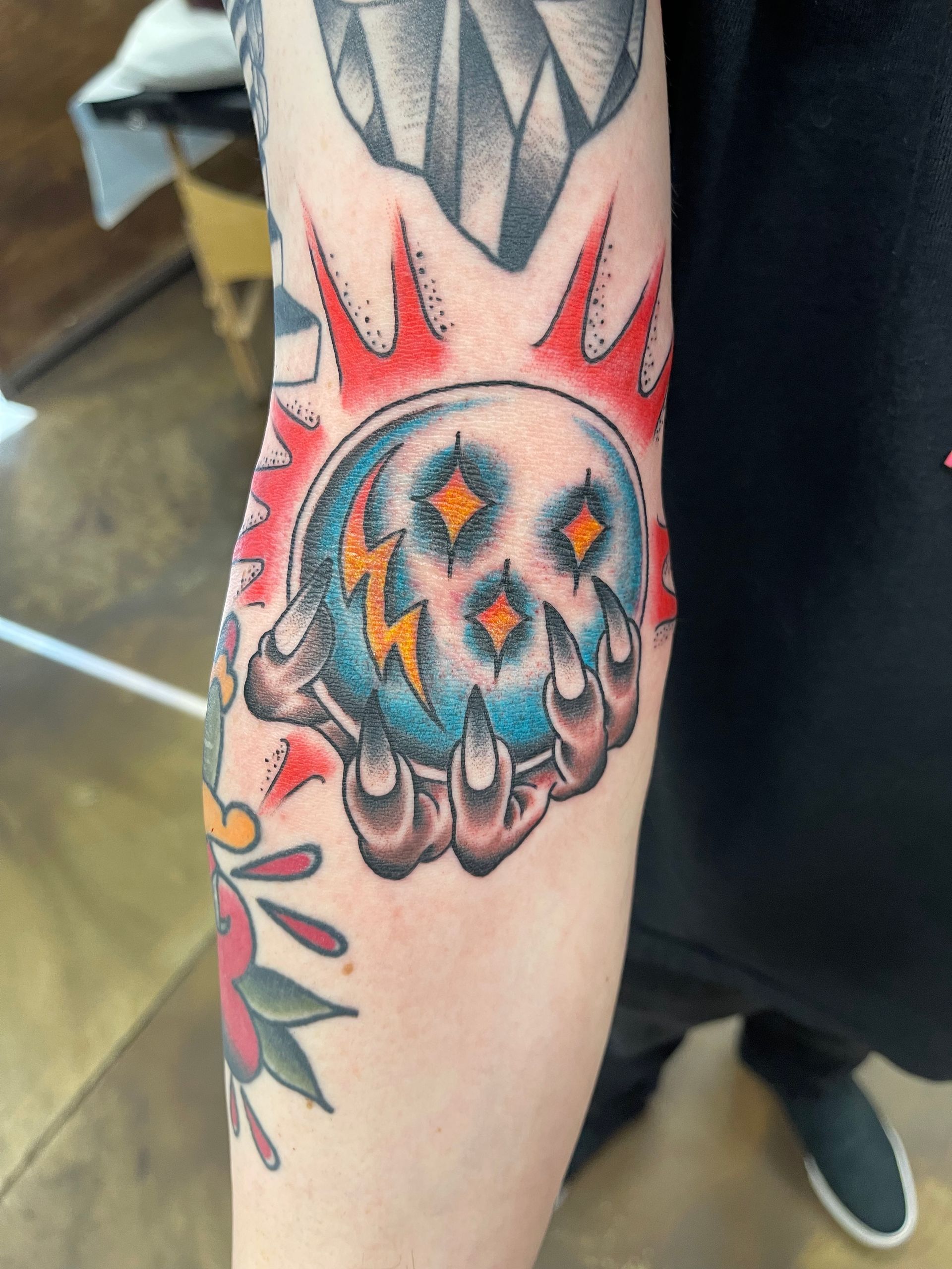 Tattoo of a hand clutching a blue crystal ball with lightning, stars, and surrounded by red and yellow bursts.