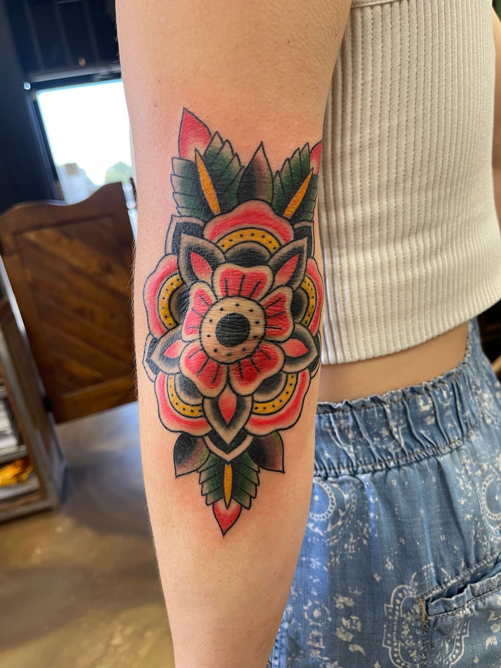 Tattoo of a red and yellow flower with green leaves on a person's elbow.
