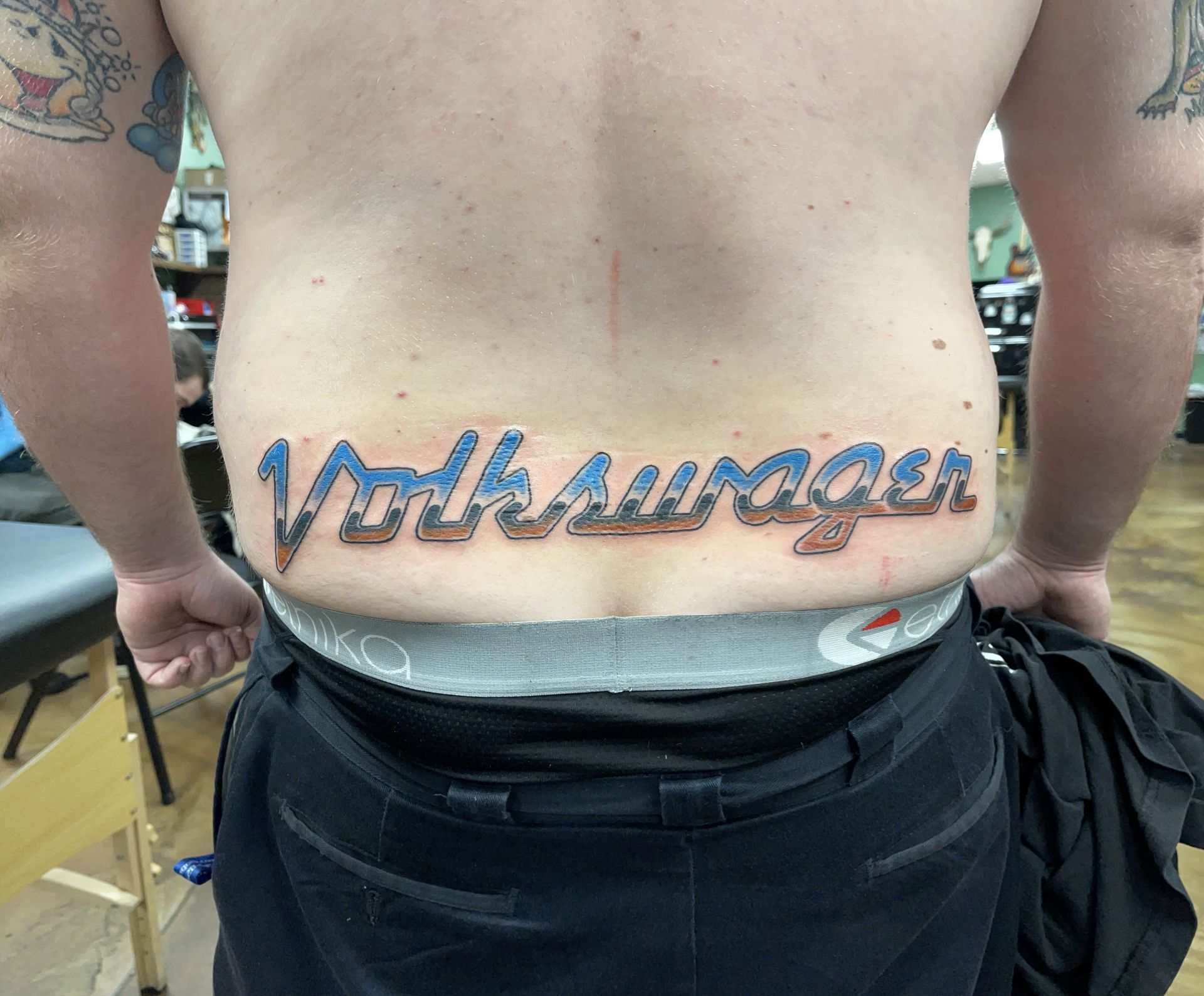 Man's lower back tattooed with the Volkswagen logo in blue and red. He wears black pants and grey underwear.