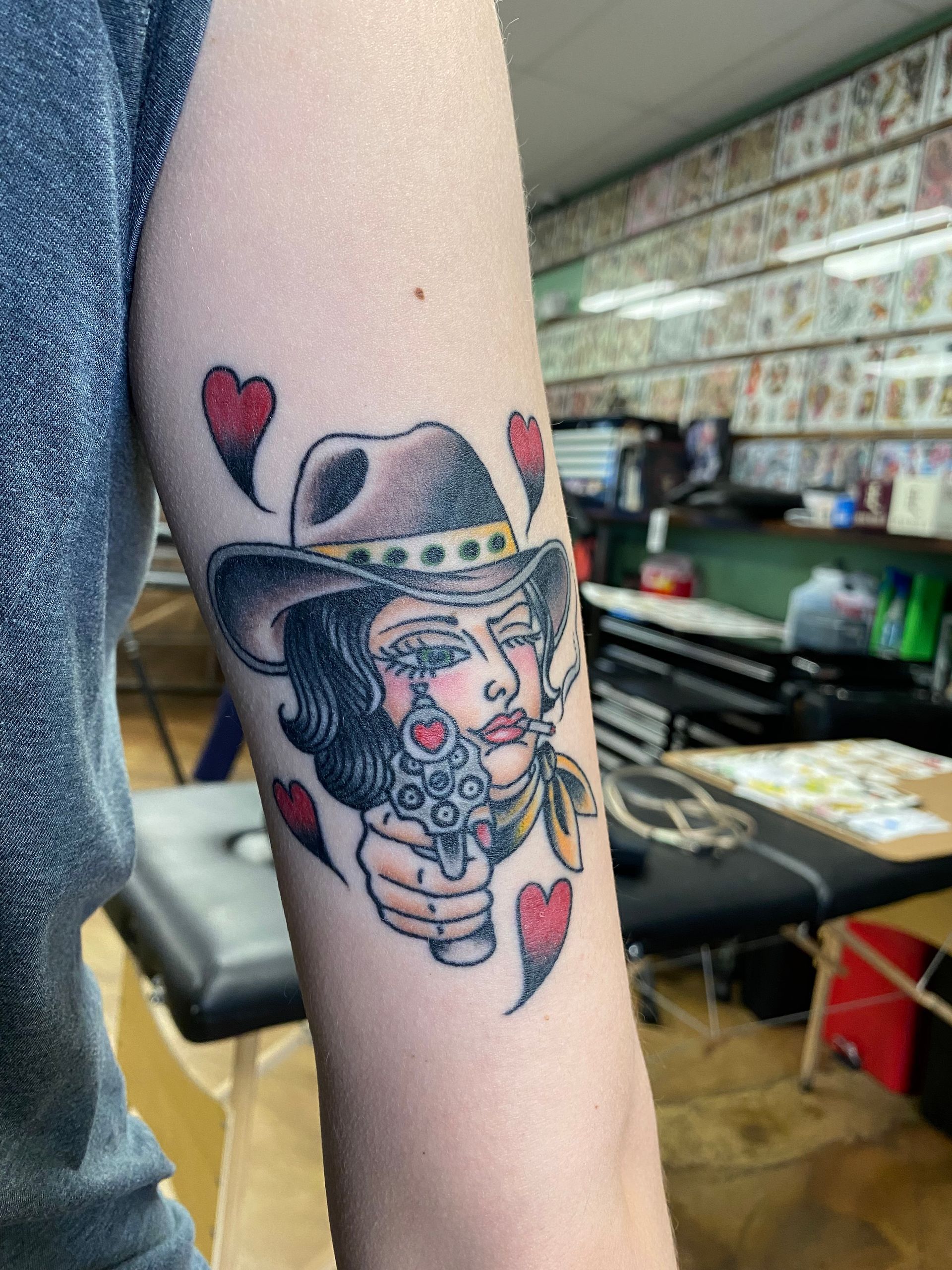 Tattoo of a cowgirl holding a gun, surrounded by hearts, on a person's arm, in a tattoo shop.