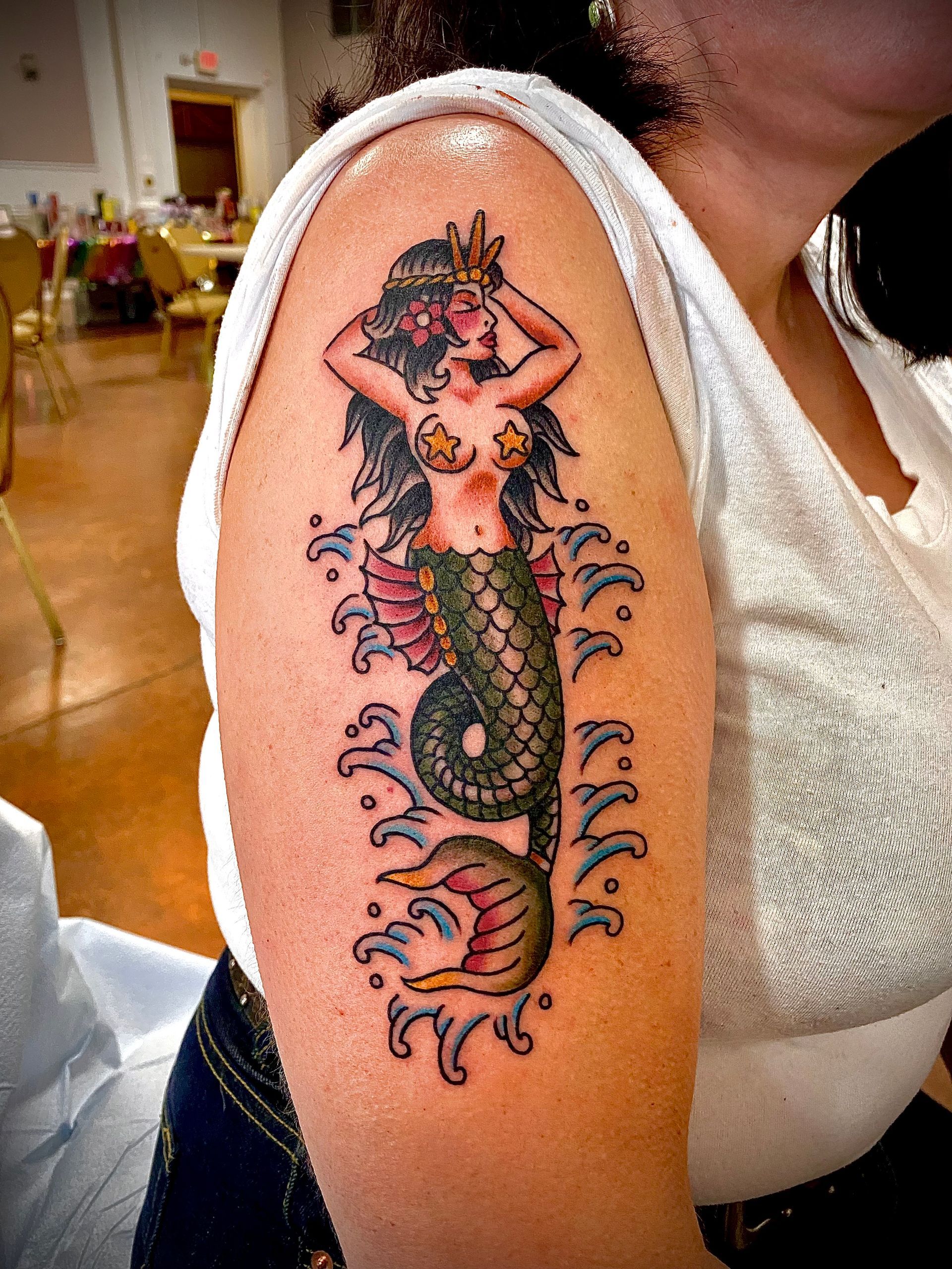Tattoo of a mermaid on a person's upper arm. The mermaid has green scales, is surrounded by blue waves and wears a crown.