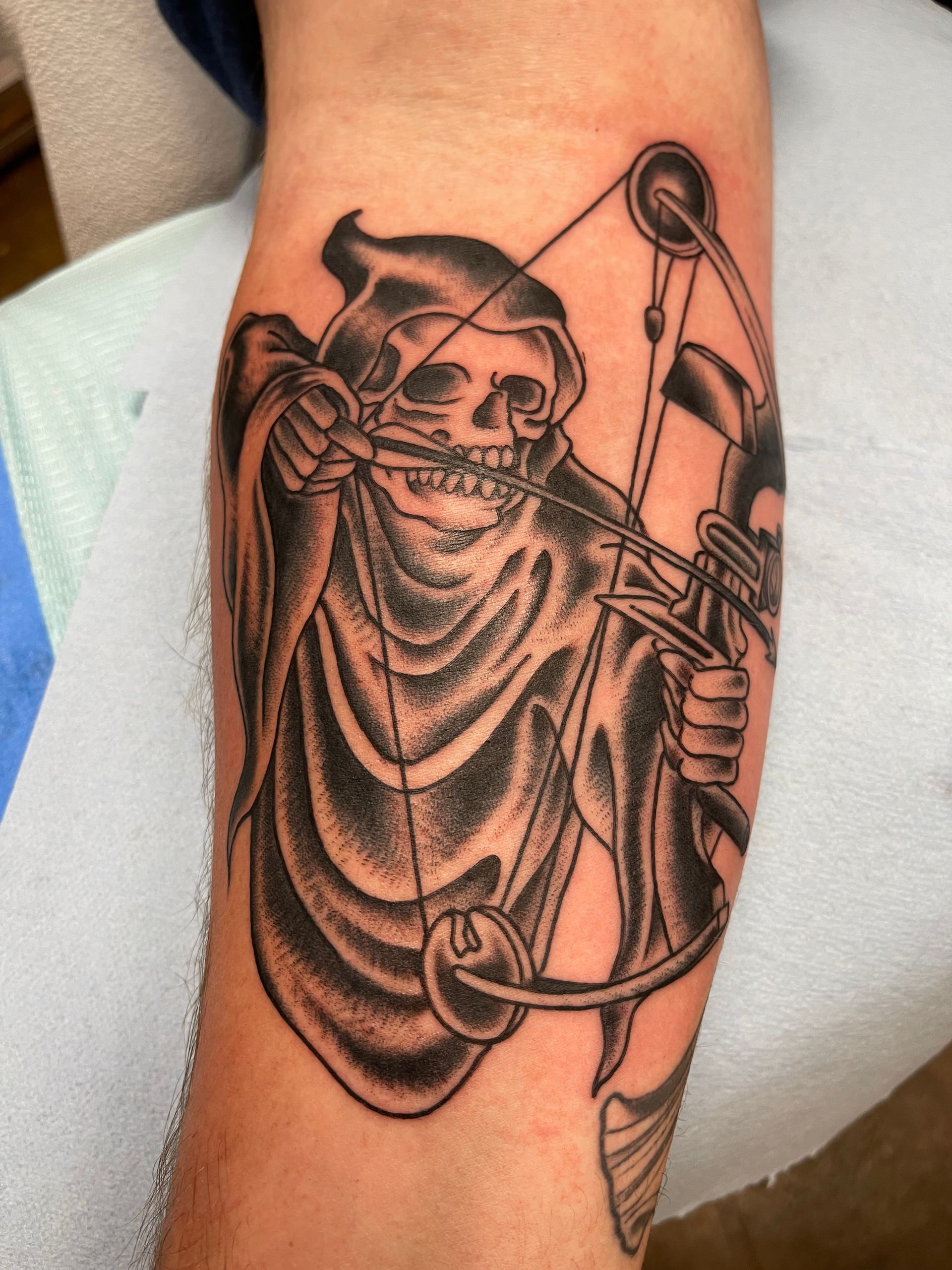 Tattoo of Grim Reaper aiming a bow. Black and gray shading on forearm.