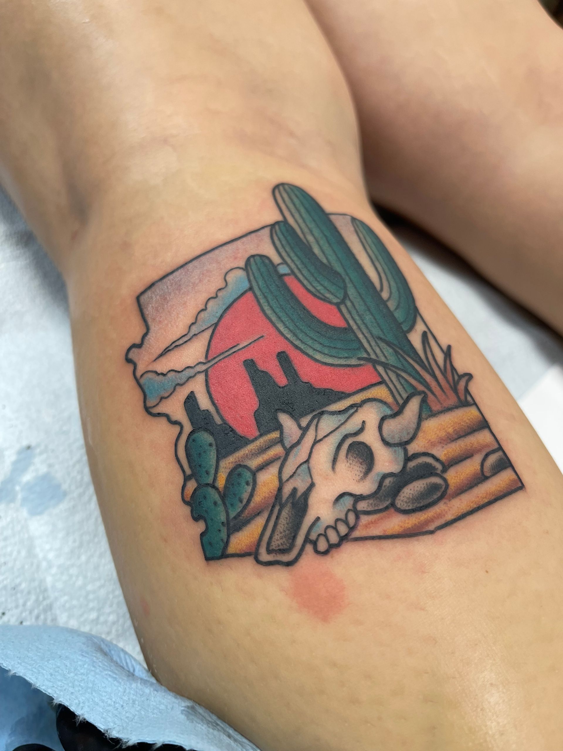 Tattoo of desert scene: cactus, skull, sun, within a stylized frame on a leg.