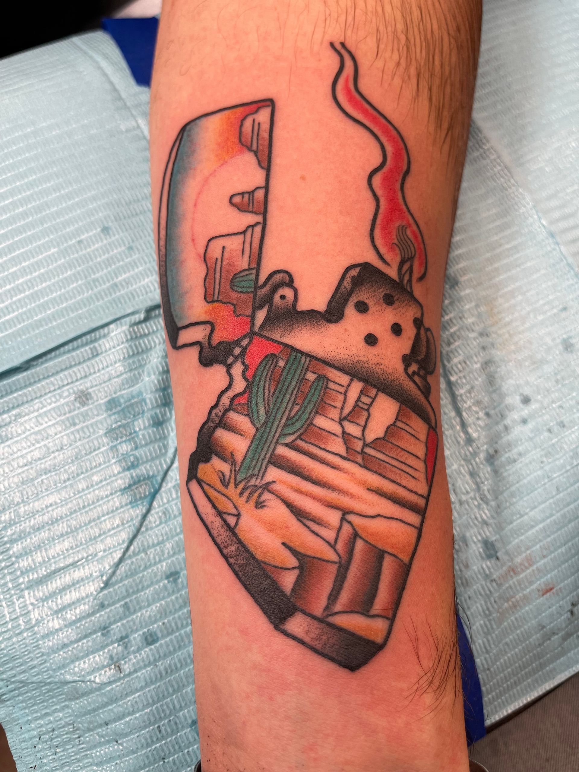 Tattoo of a desert scene inside an open lighter, with a flame rising. Black, red, blue, and tan colors on arm.