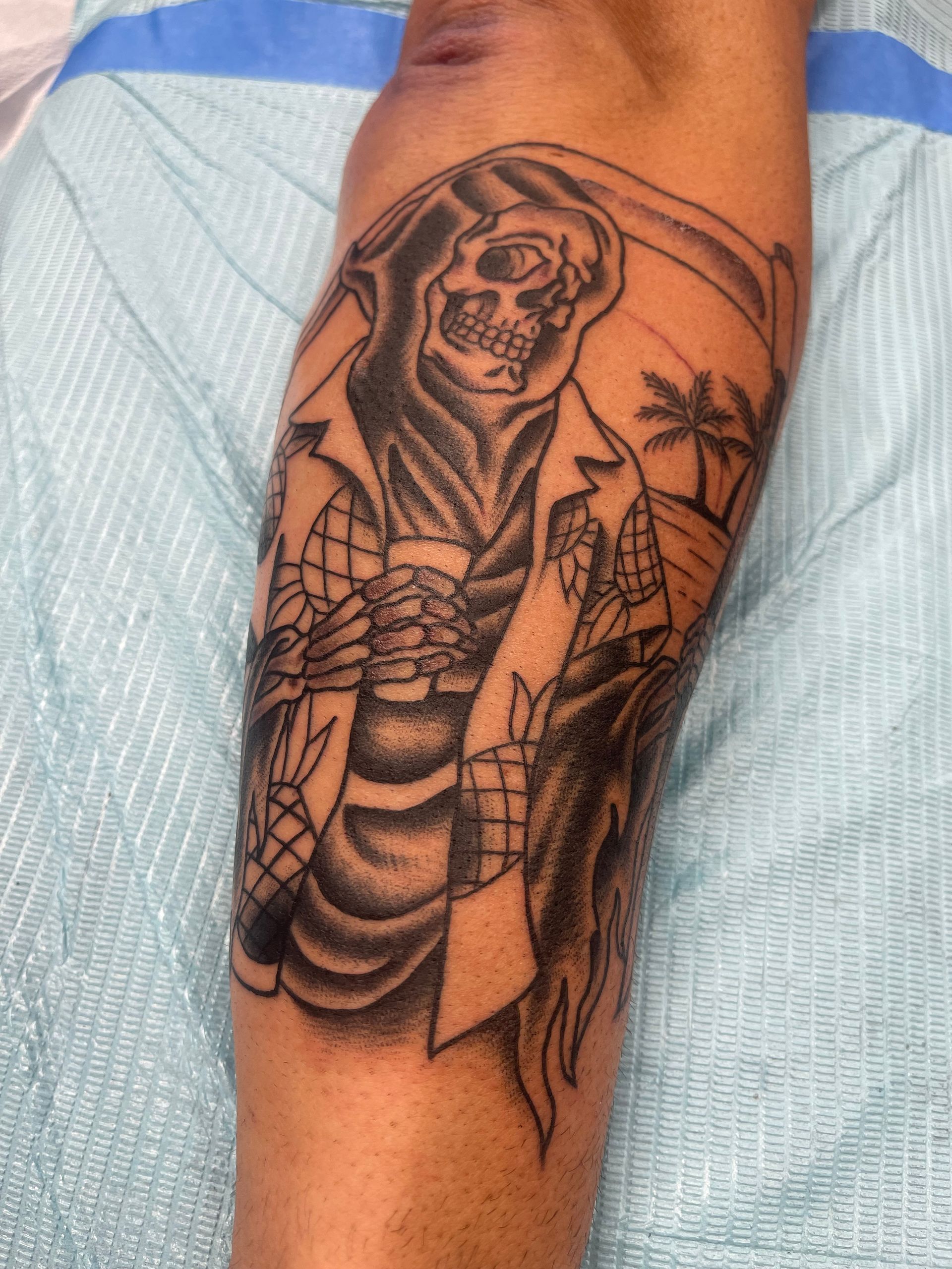 Tattoo on arm depicts the Grim Reaper holding a drink, wearing a Hawaiian shirt with a tropical scene in the background.