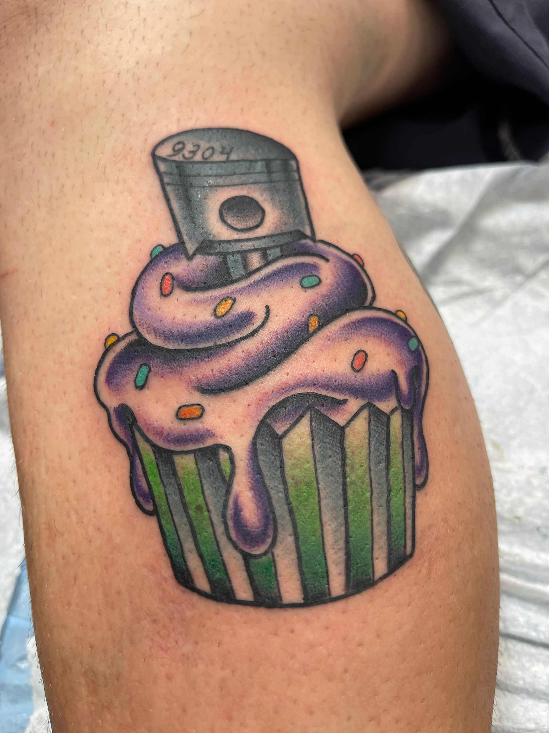 Tattoo of a cupcake with purple icing, sprinkles, and a piston on top. Green and grey striped wrapper.