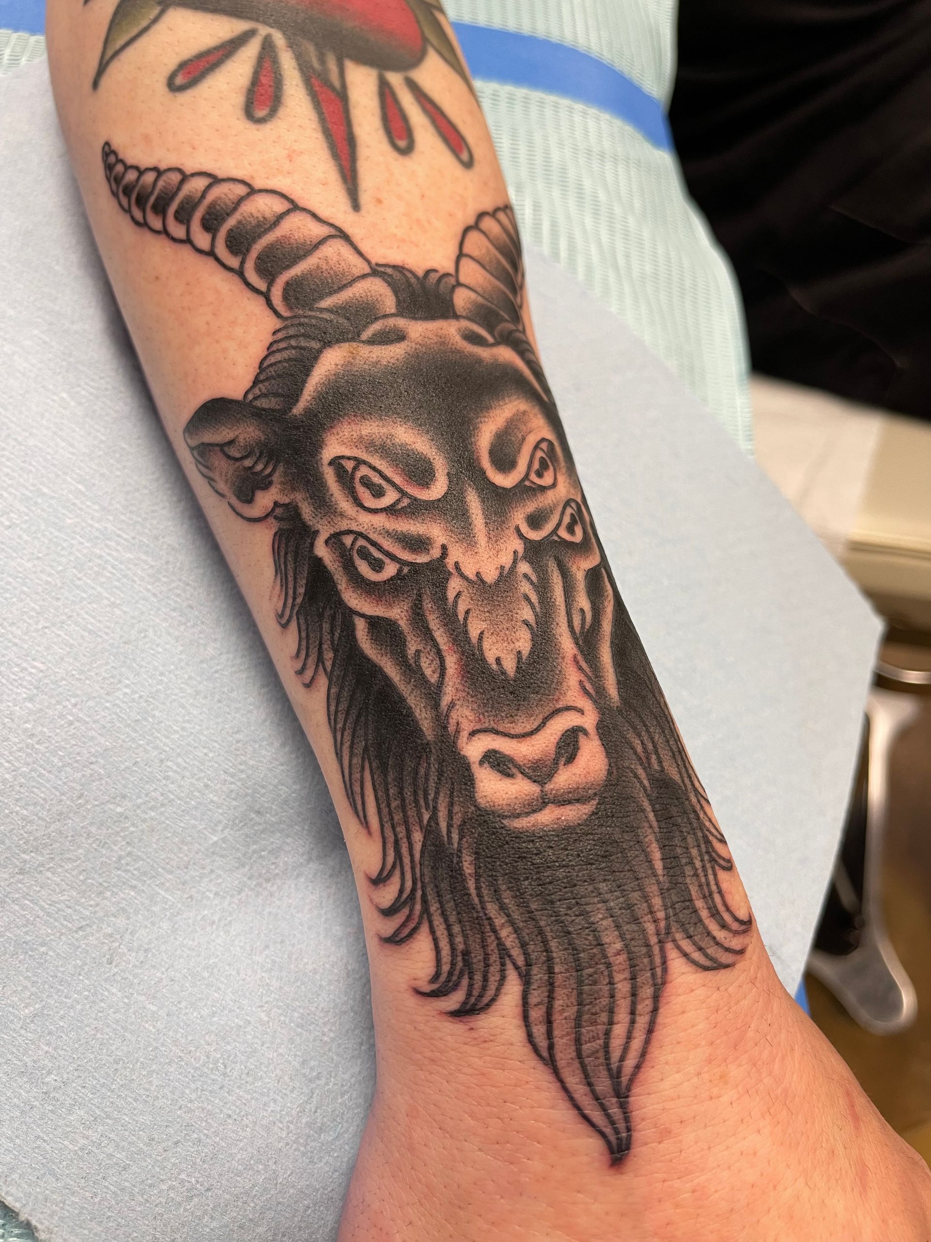Tattoo of a goat with multiple eyes and large horns on a person's arm. Black and gray shading.