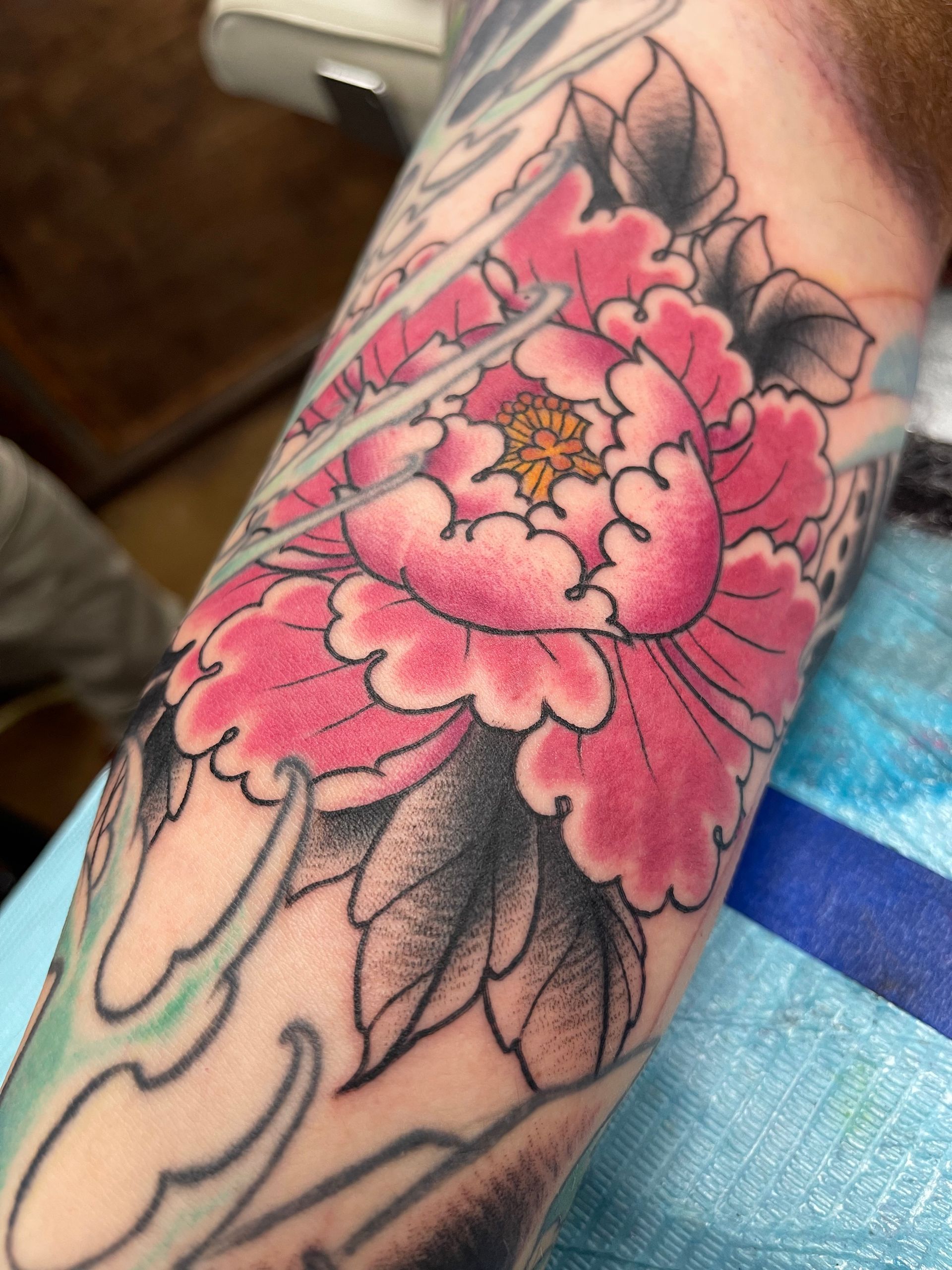Pink peony tattoo on arm with black and gray leaves.