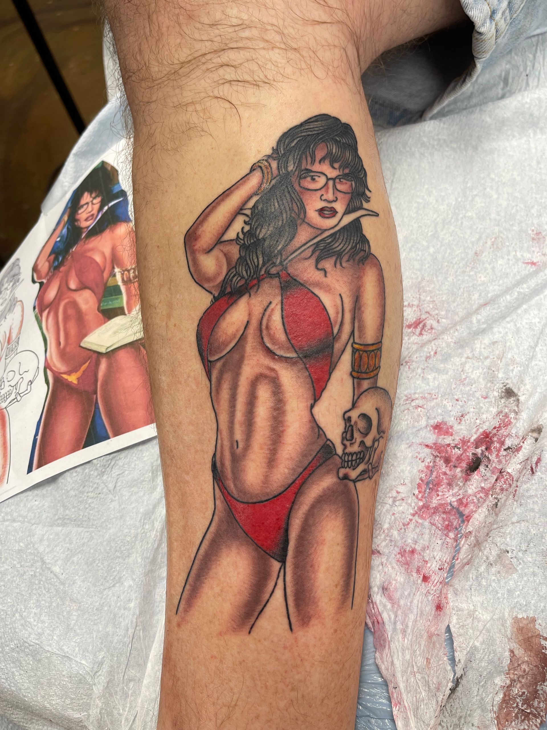 Tattoo of a woman in red lingerie holding a skull, on a leg. The tattoo is next to the original image.