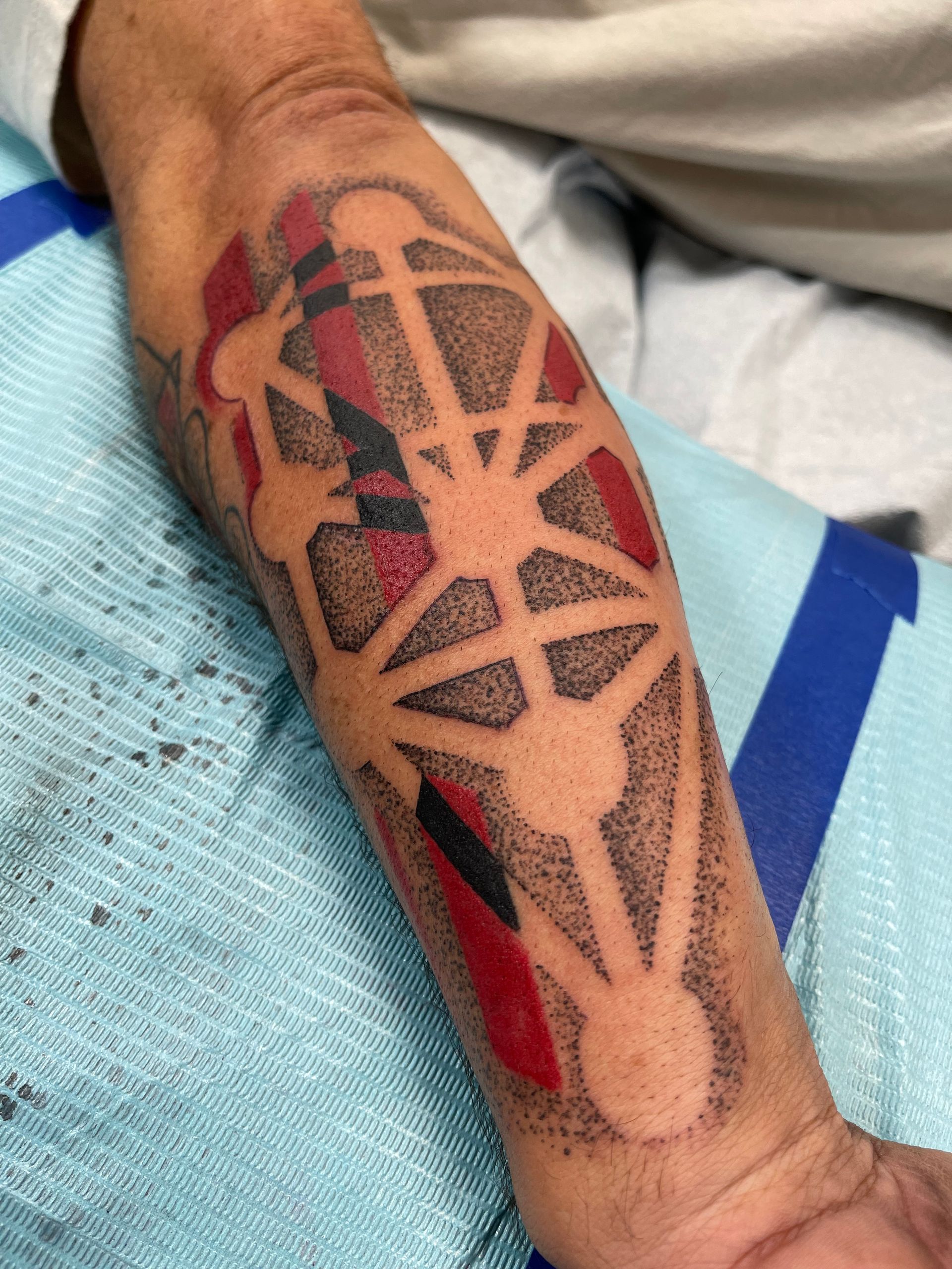 Arm with a tattoo in red, black, and tan, featuring a geometric Tree of Life design.