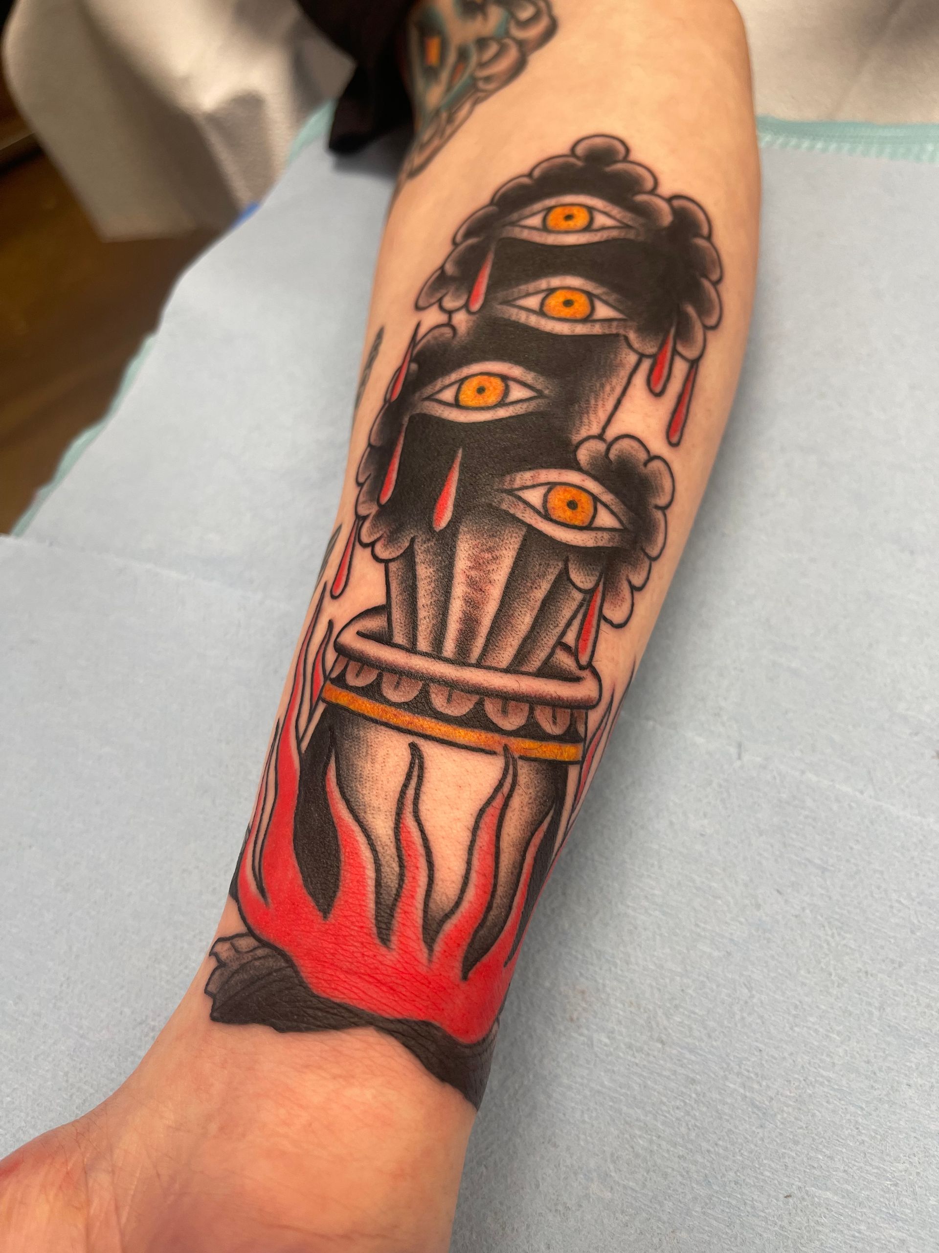 Tattoo of a black cauldron with flames, topped by three eye-filled, cloud-like shapes on a person's arm.