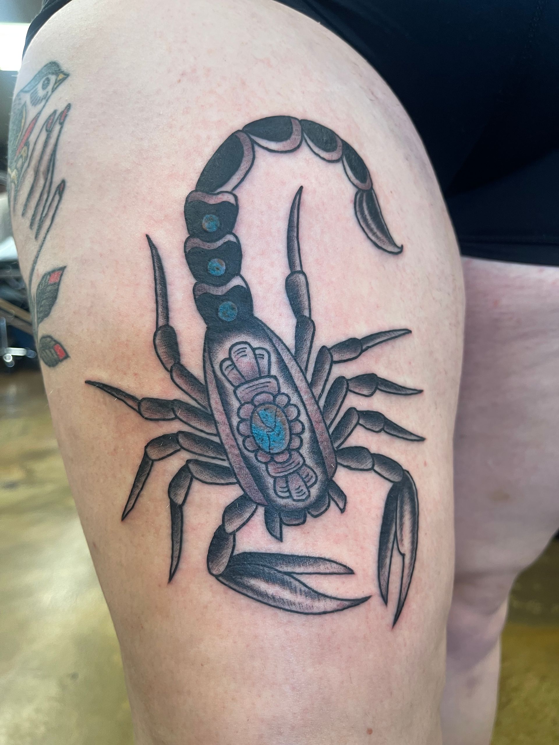 Black and gray scorpion tattoo on a person's thigh, with teal and blue accents.