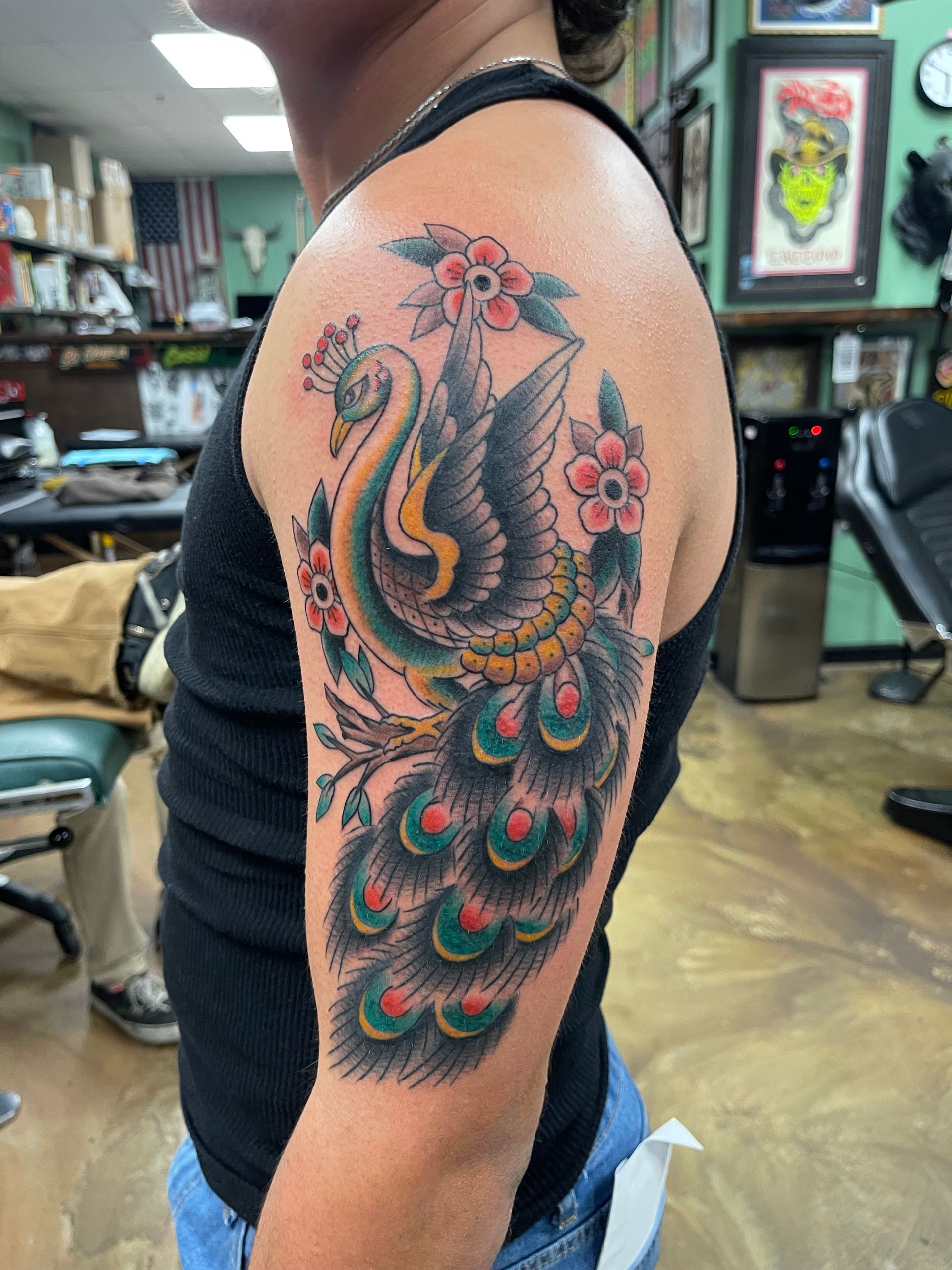 Tattoo of a peacock with colorful feathers on a person's upper arm, with a tattoo parlor background.