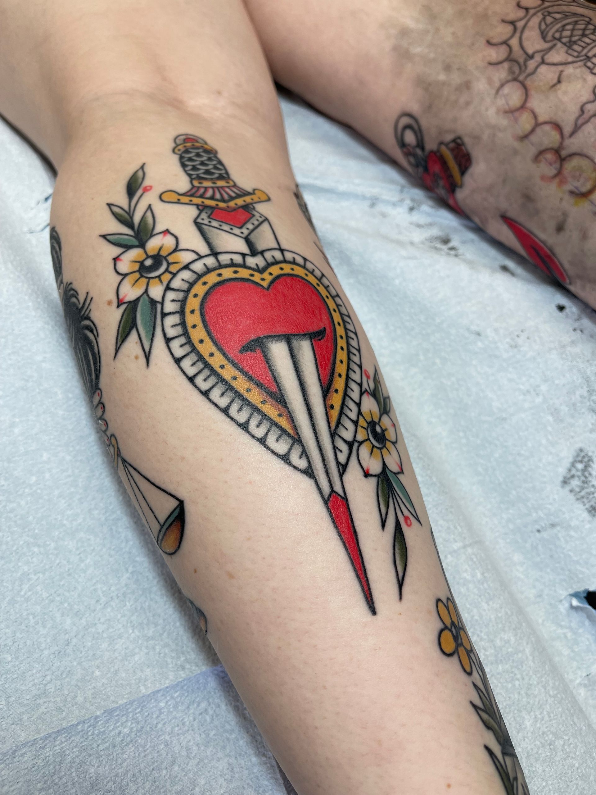 Tattoo of a heart pierced by a dagger, with flowers and leaves on a person's arm.