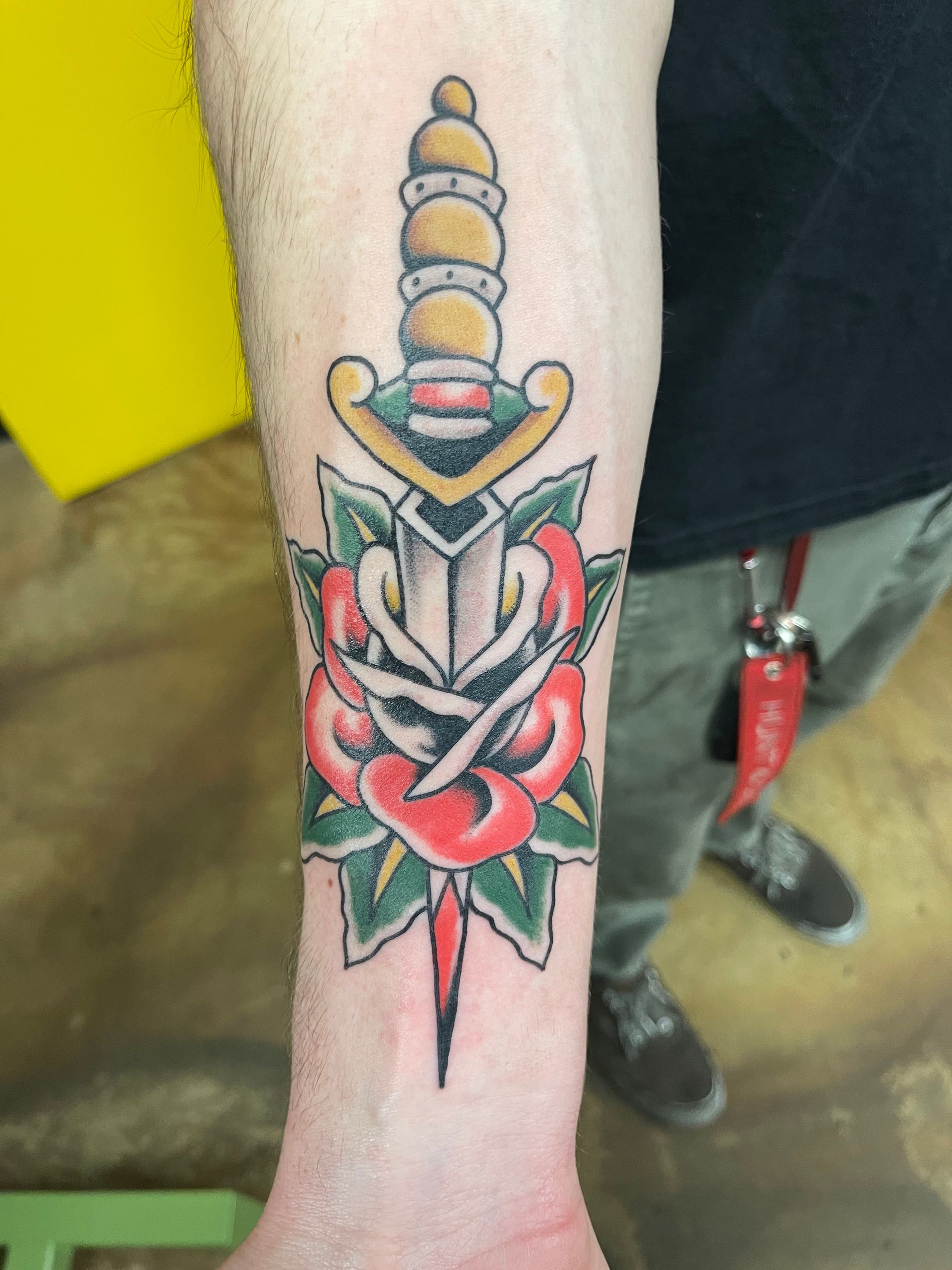 Tattoo of a dagger through a red rose on a person's forearm. Green leaves and a yellow background.