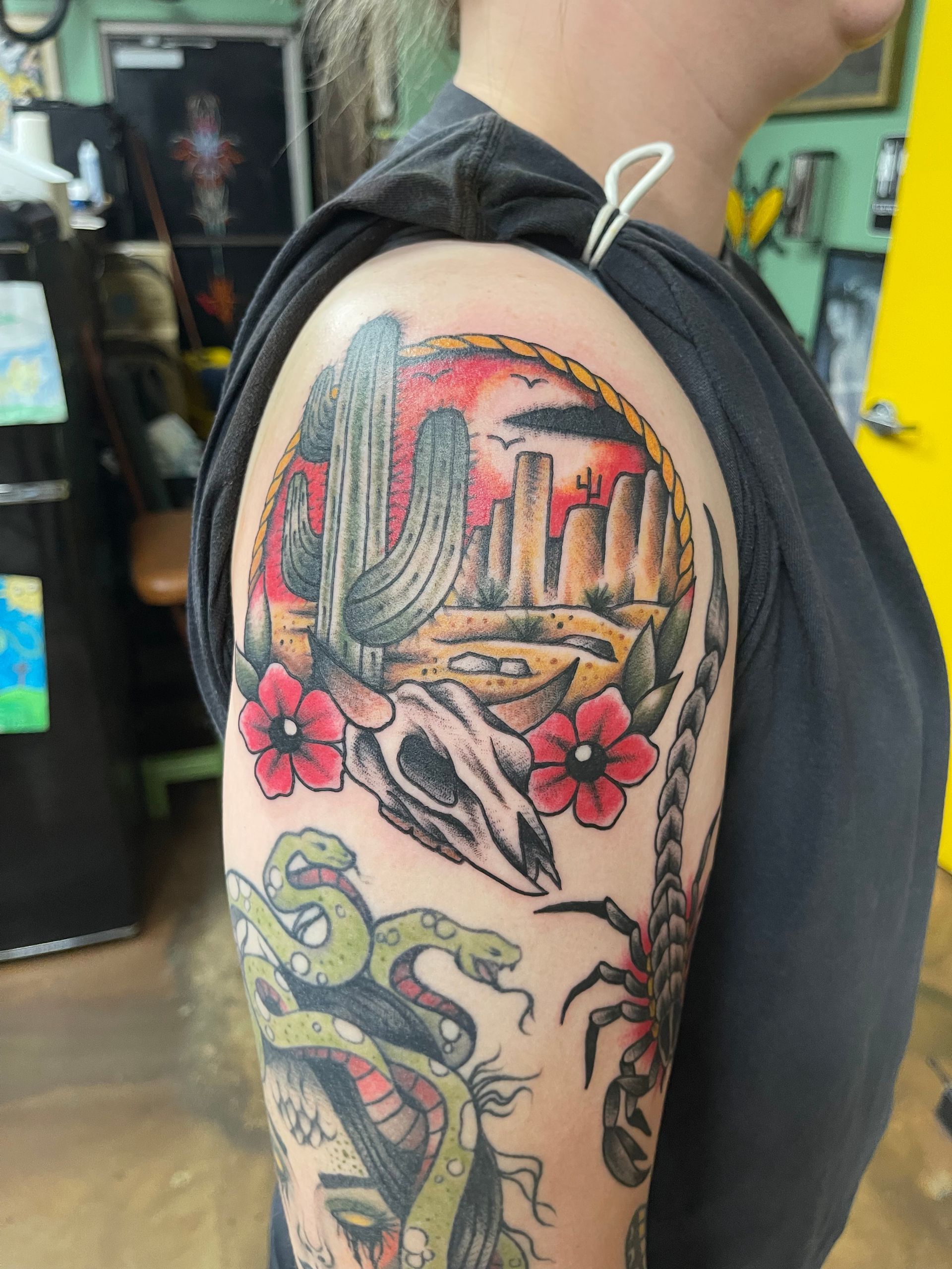 Tattoo of a desert scene with cacti, skull, flowers, and snakes on a person's arm.