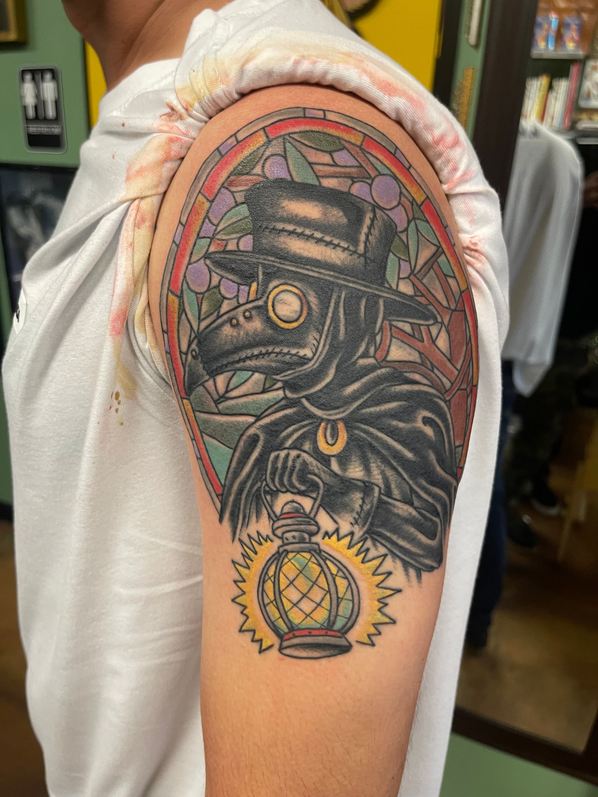 Tattoo of a plague doctor in stained glass on a person's upper arm, holding a lantern.