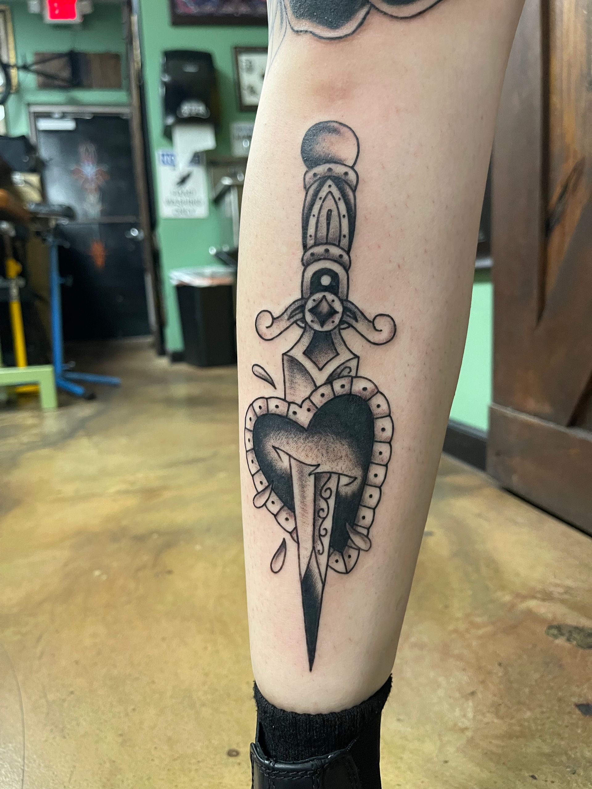 Tattoo of a dagger piercing a heart with a beaded border, on a leg. The tattoo is black and grey.