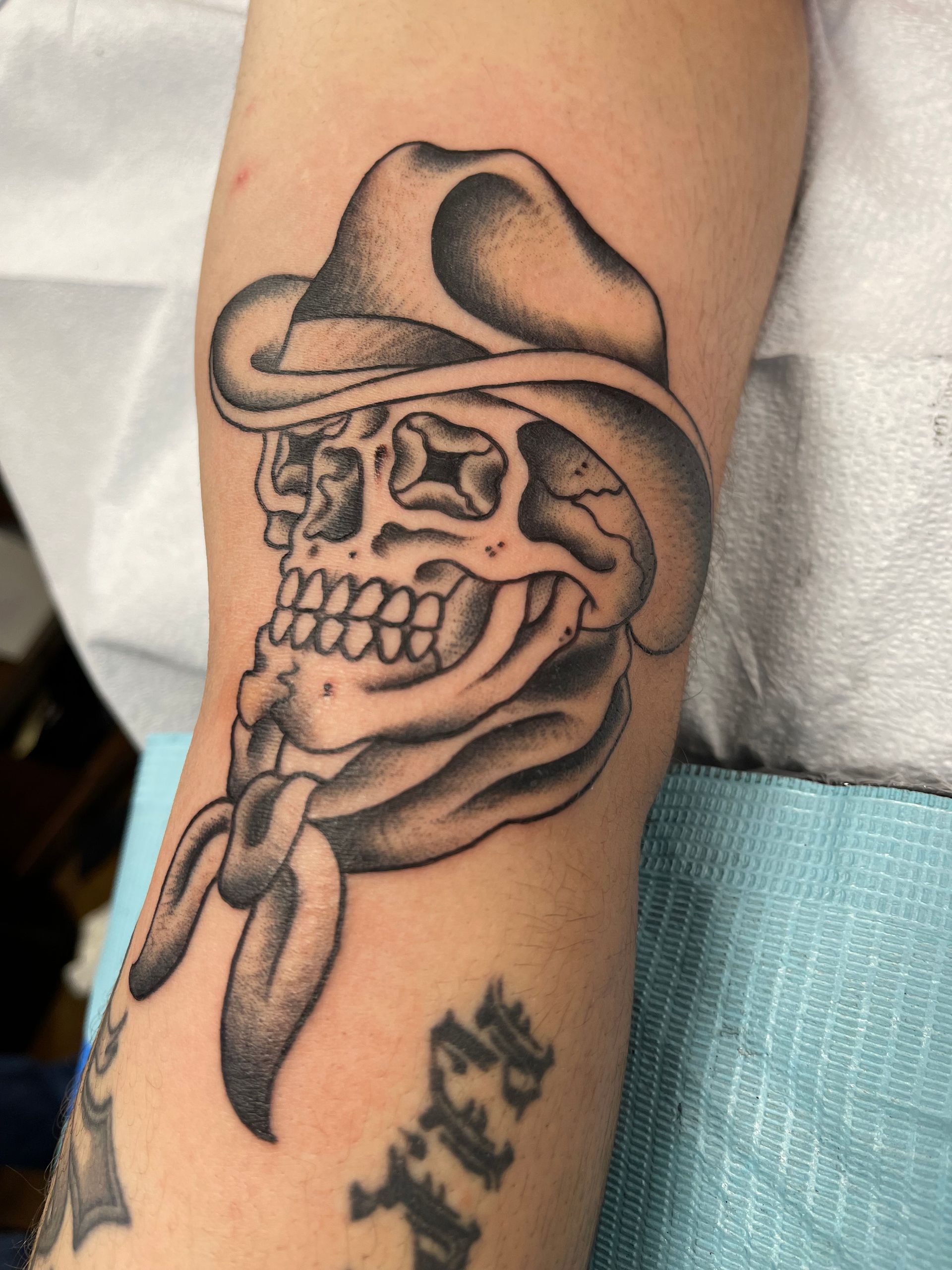 Tattoo of a skull wearing a cowboy hat and bandana. Black and gray shading on forearm.