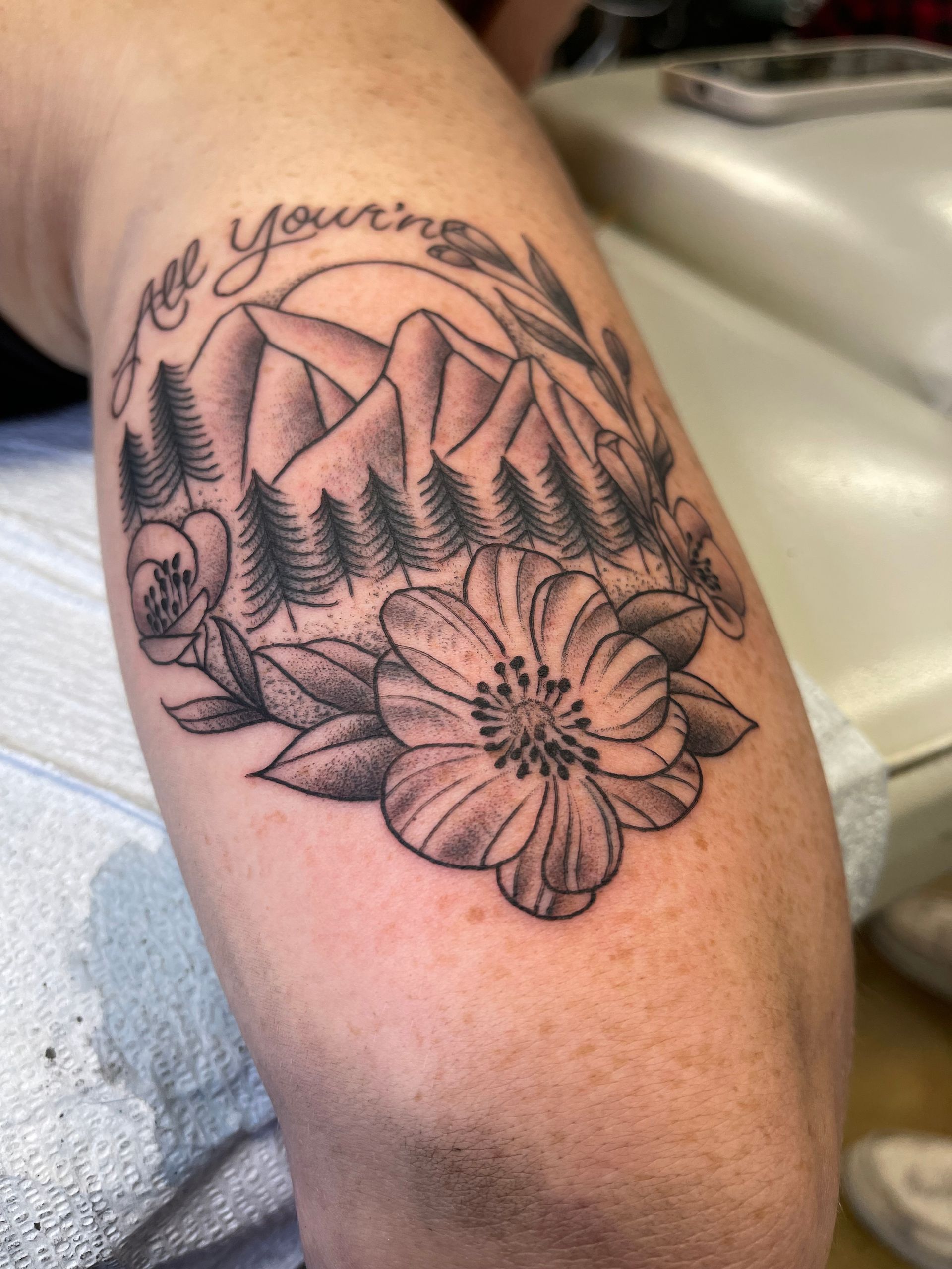 Tattoo on arm features mountains, trees, flower, foliage, and text 