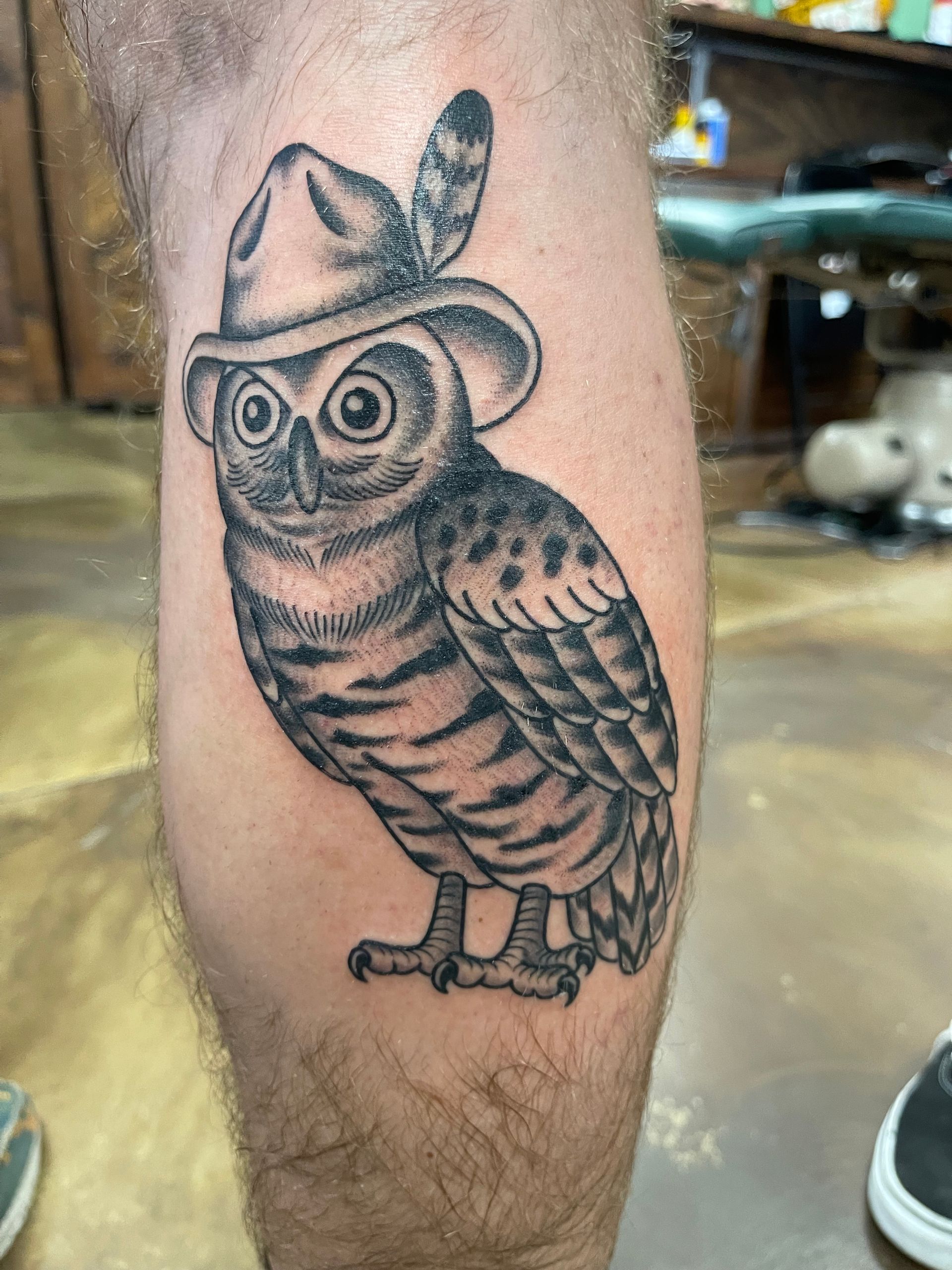 Tattoo of an owl wearing a hat with a feather on a leg. Black and gray, detailed shading.