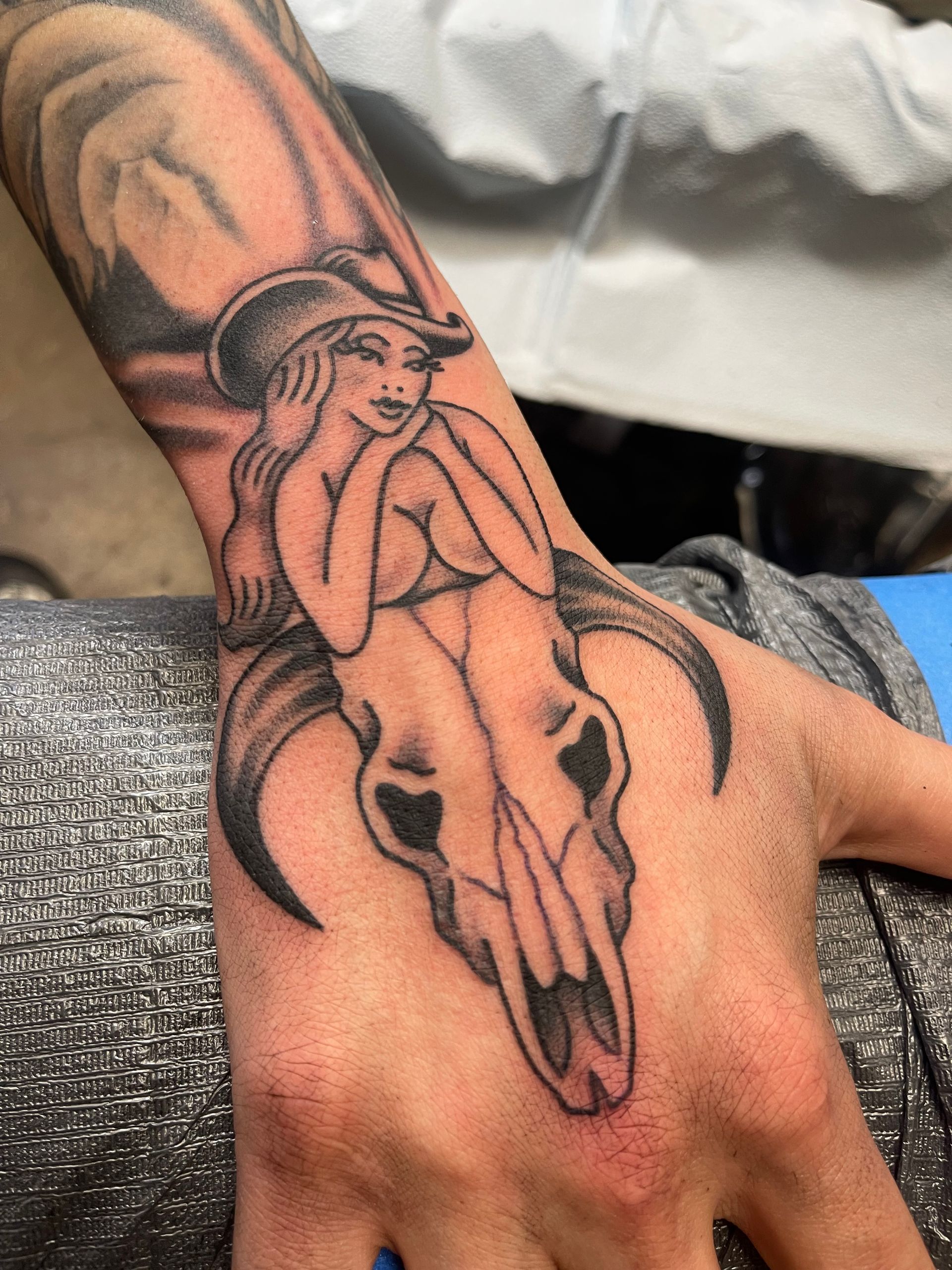 Tattoo on hand: cow skull with woman in cowboy hat posed over it in black and gray.