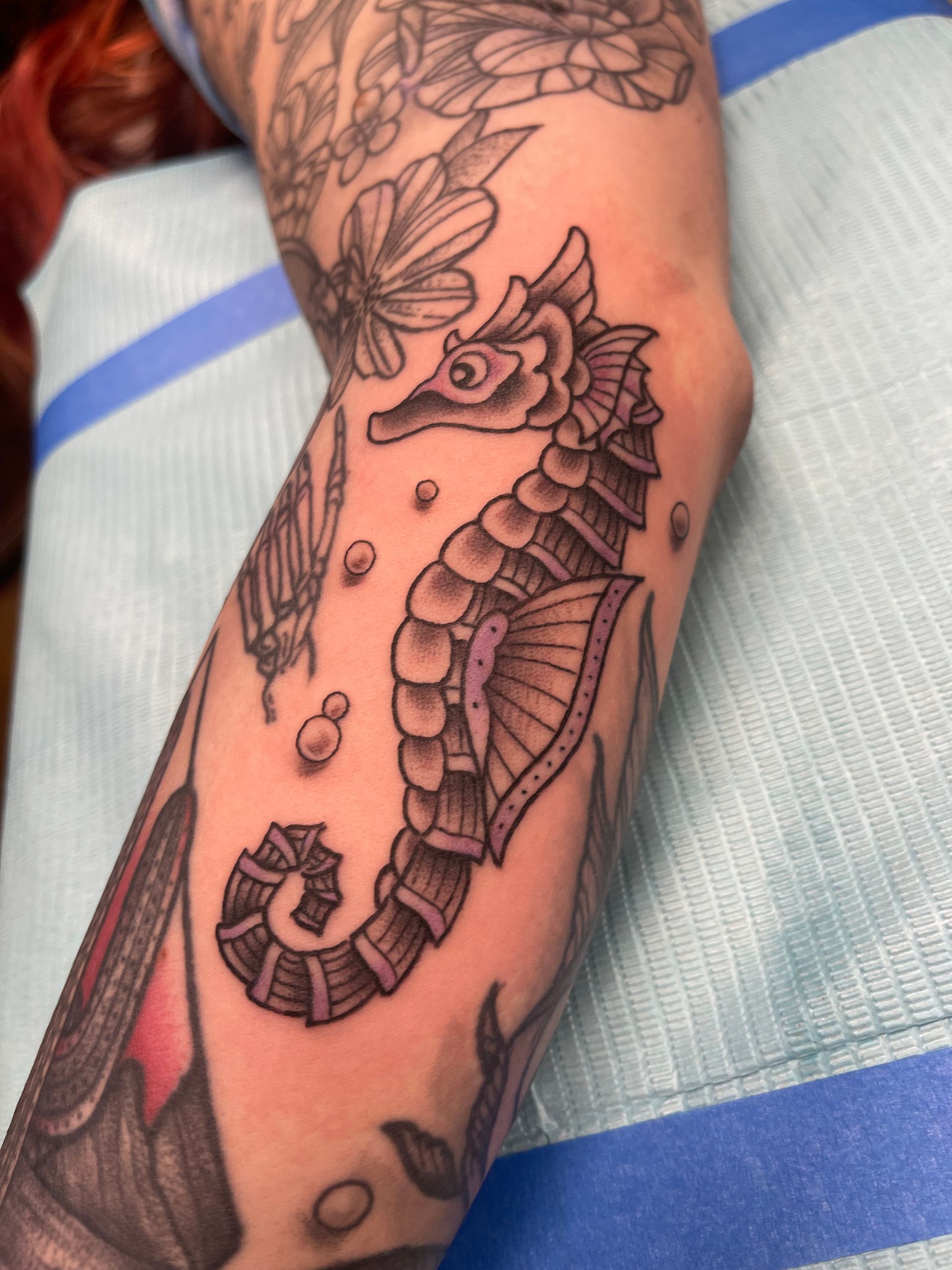 Black and white seahorse tattoo on a person's arm, surrounded by other tattoos and bubbles.