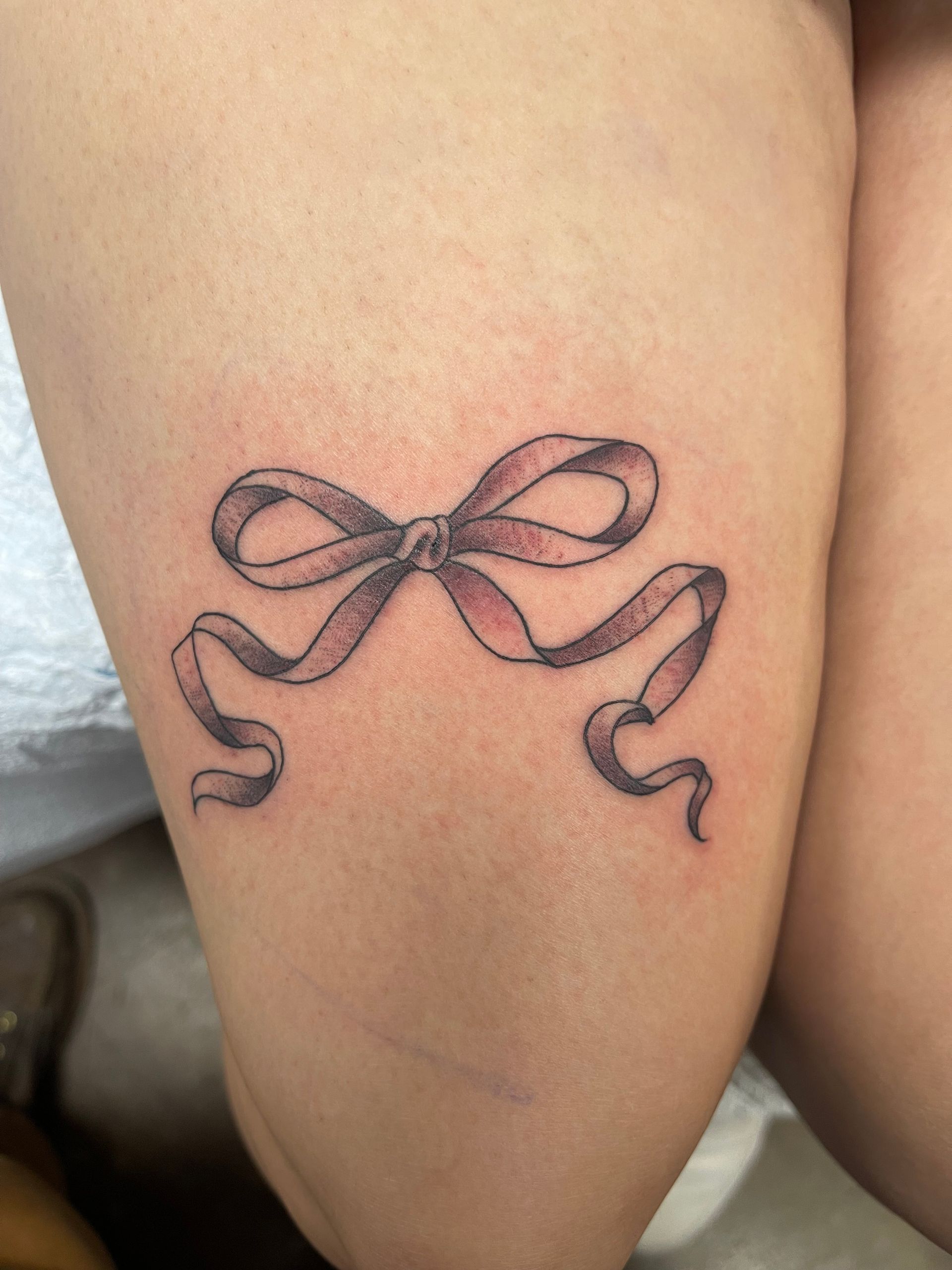 A bow tattoo on the upper thigh with black outline and subtle pink shading.