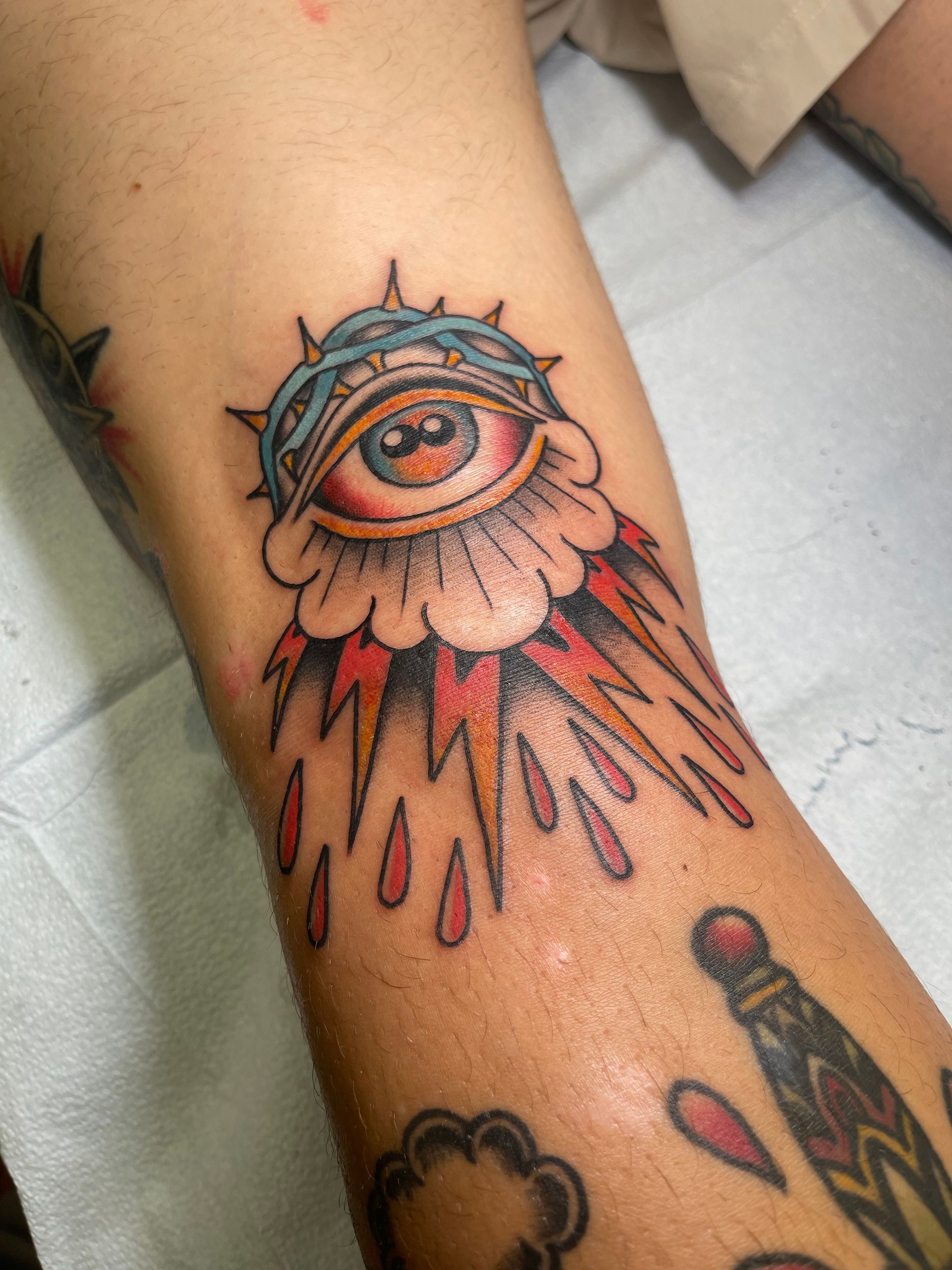 Tattoo of a stylized eye with a crown of thorns and radiating rays on an arm. Red, blue, orange.