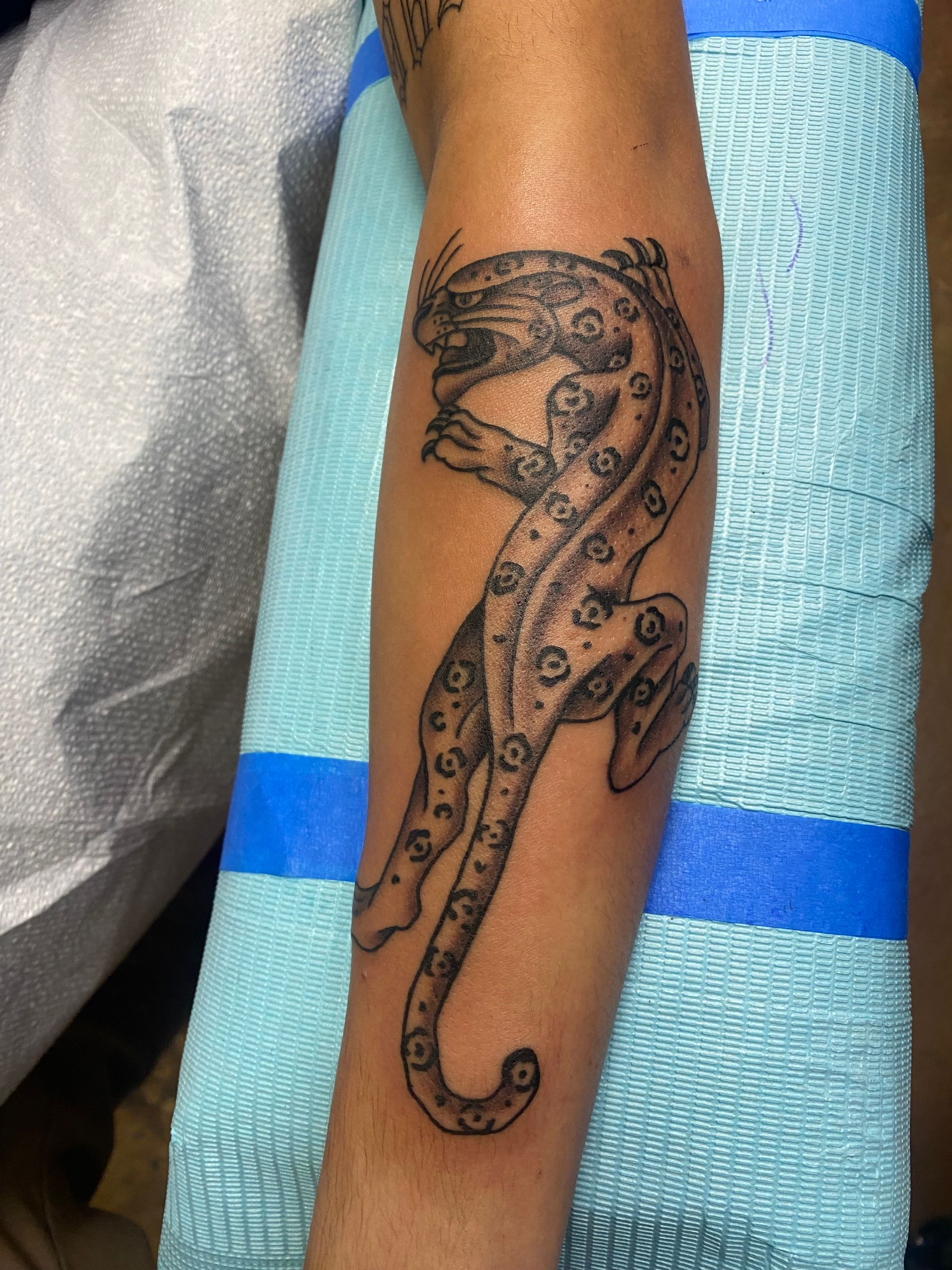 Black and gray tattoo of a leopard on a forearm; the animal is depicted with spots, in a leaping position.