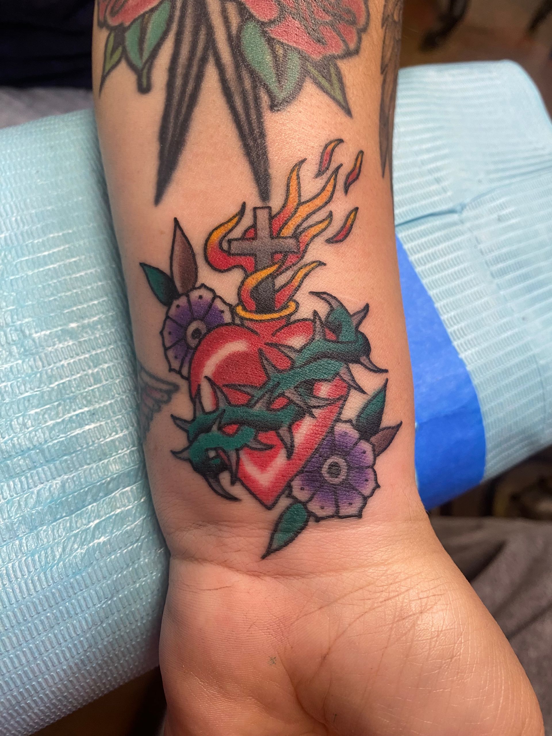 Tattoo of a heart with flames, a cross, and a crown of thorns on a wrist surrounded by flowers.