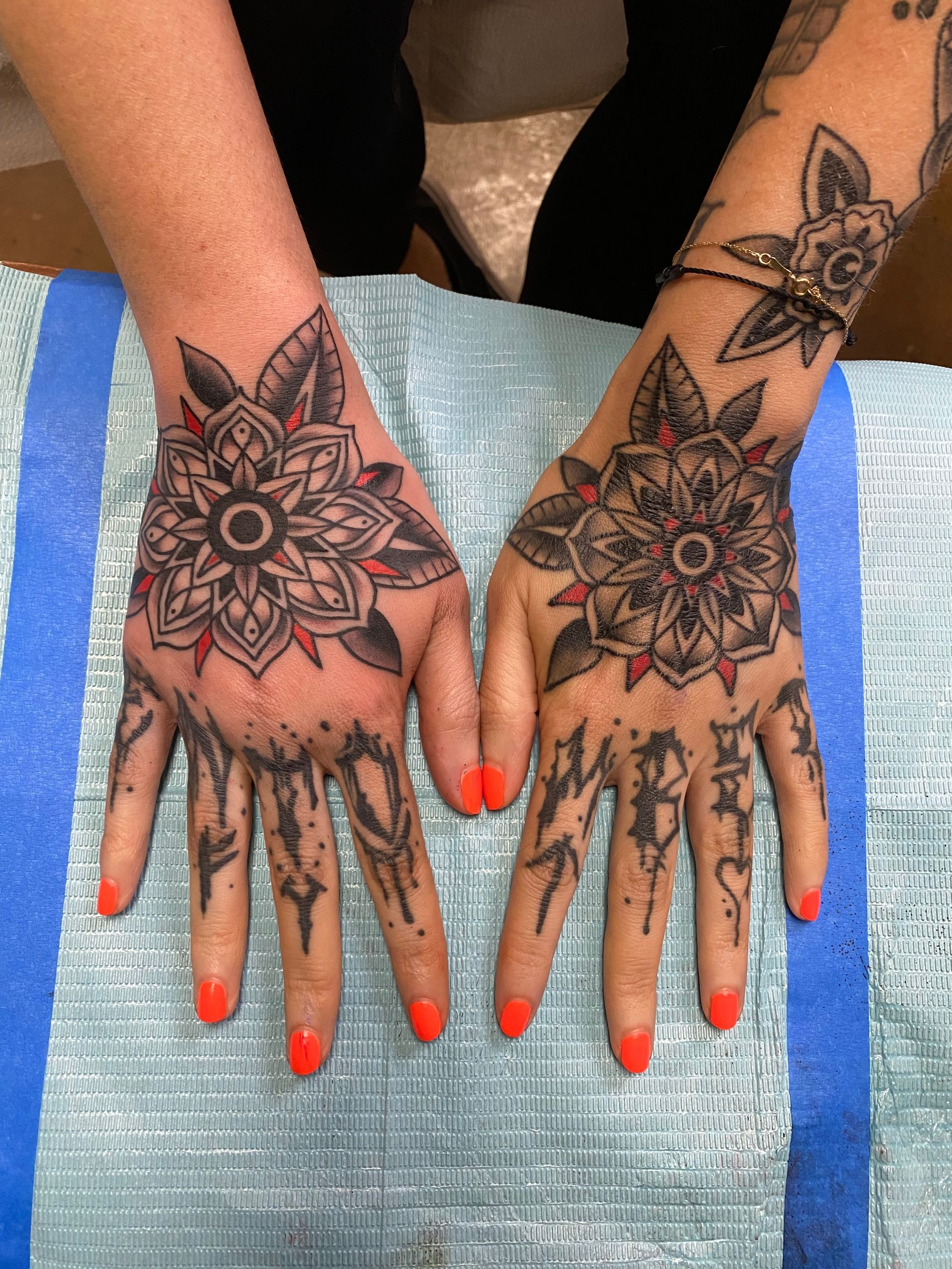 Hands with elaborate black and red mandala tattoos on palms and finger tattoos, orange nail polish.