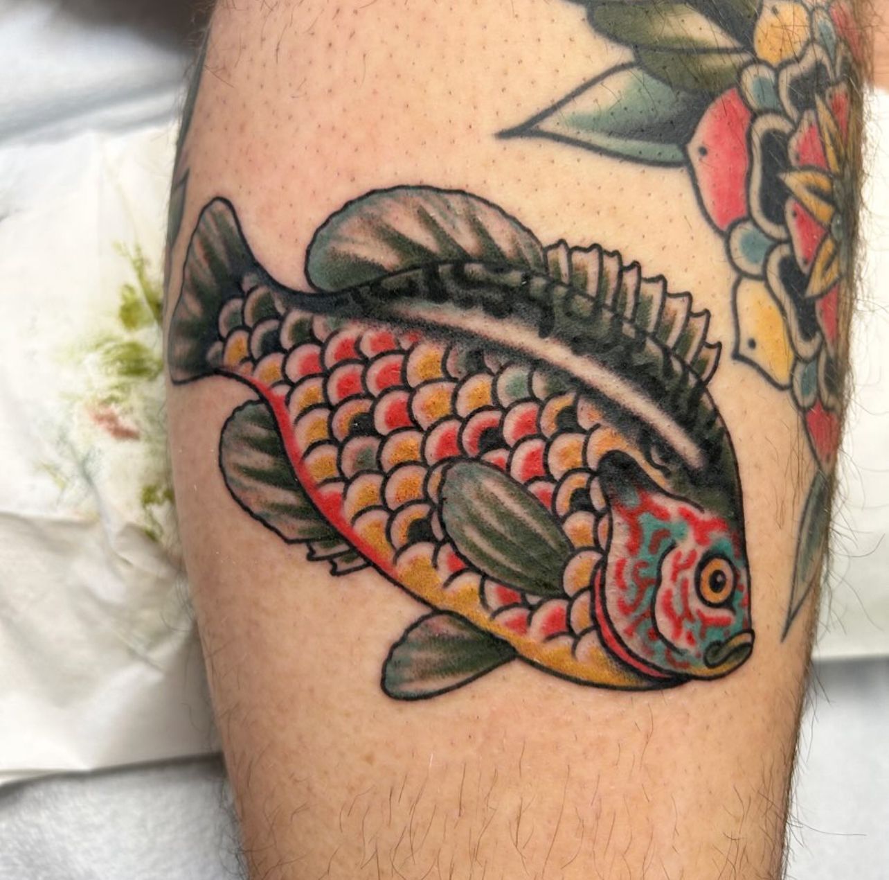 Colorful fish tattoo on a leg, scales in red and yellow, with black and green accents.