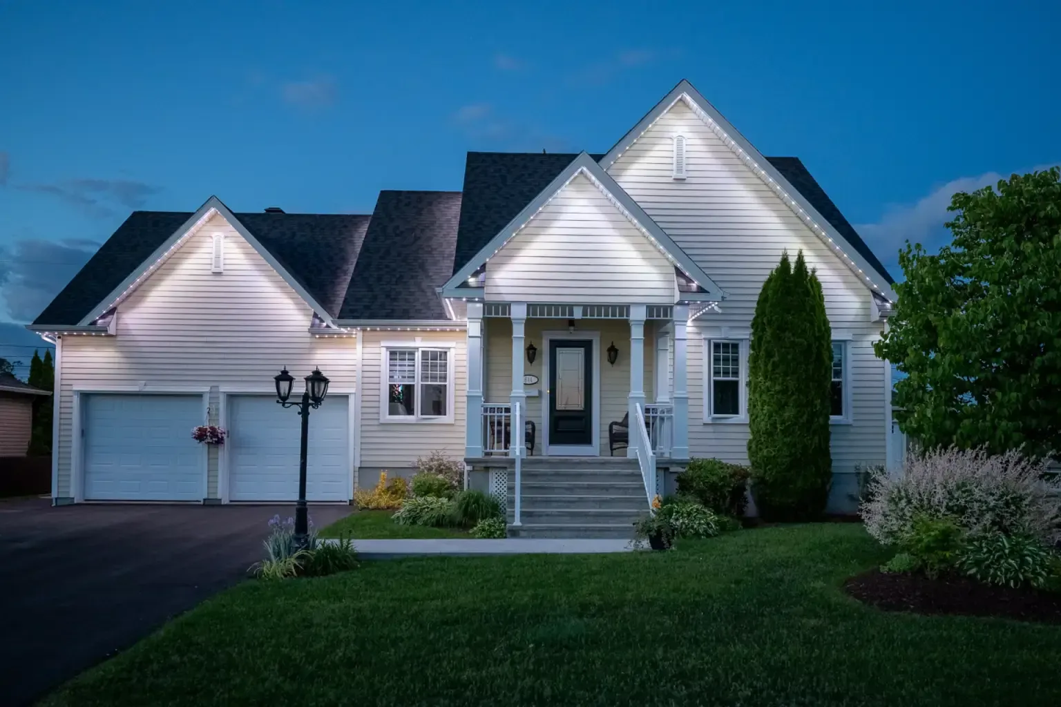 Lit-up white house with a dark roof and garage doors at twilight; green lawn and landscaping.