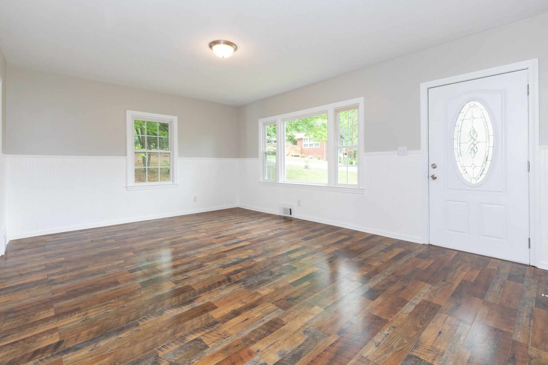 Empty living room with hardwood floors, white walls, windows, and a front door with oval glass.