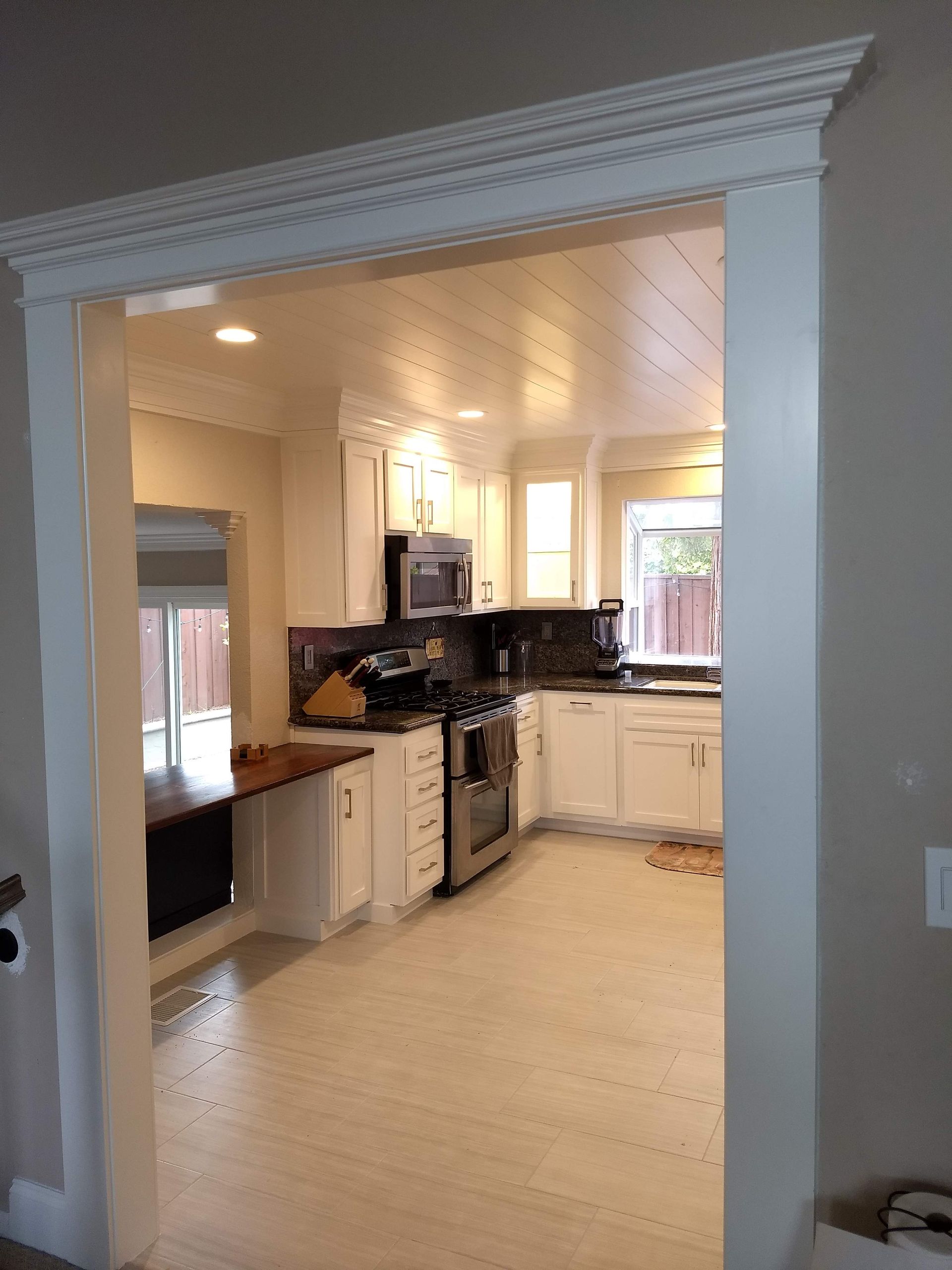 Bright kitchen viewed through a wide doorway, with white cabinets, black countertops, and beige tile floor.