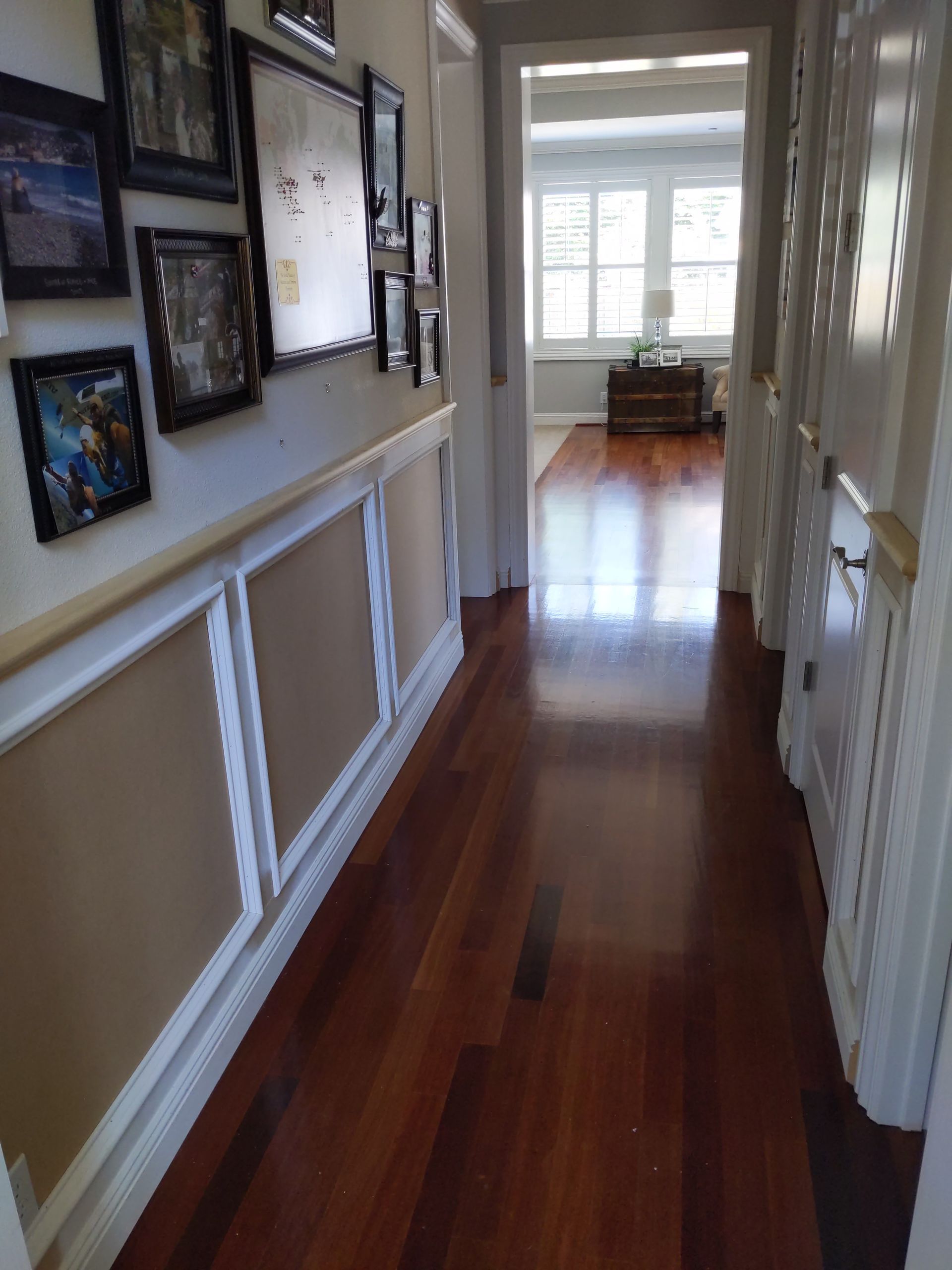 Narrow hallway with framed photos, white trim, and hardwood floor leading to a bright room with windows.