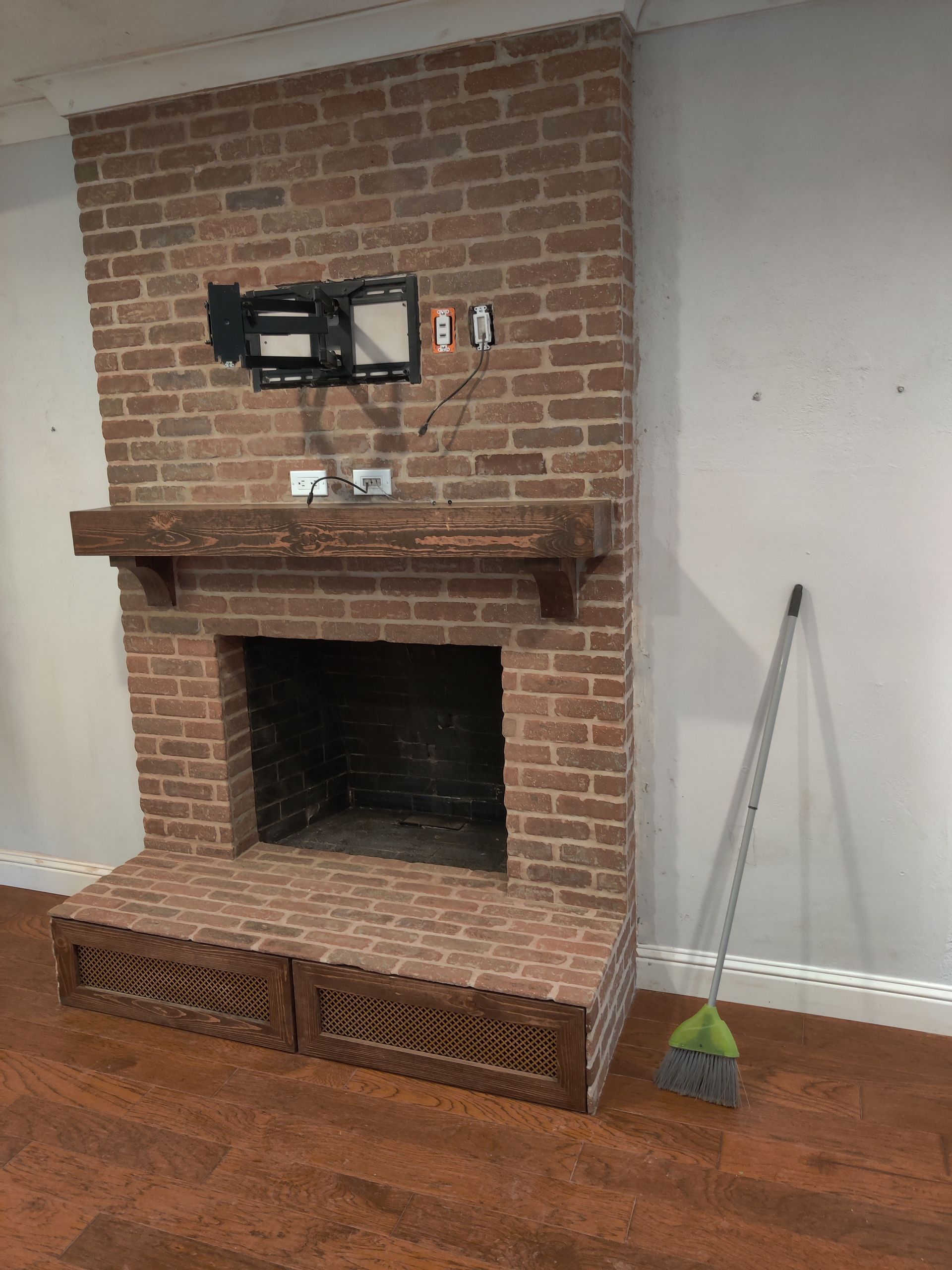 Brick fireplace with mantel and TV mount in a room, with a broom leaning nearby and hardwood floors.
