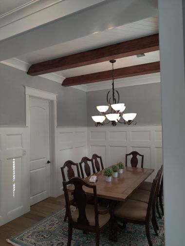 Dining room with dark wood table, eight chairs, chandelier, white wainscoting, and exposed ceiling beams.