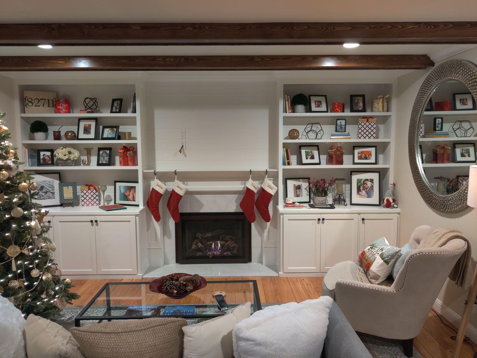 Cozy living room with a lit Christmas tree, fireplace, stockings, and built-in shelves with framed photos.