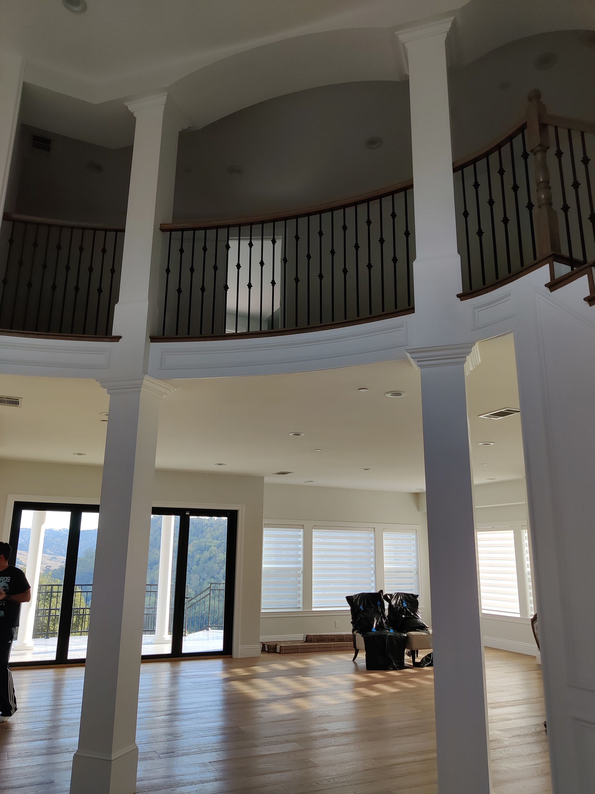 Spacious two-story living room with balcony railing, white columns, tiled floor, and bright glass doors.