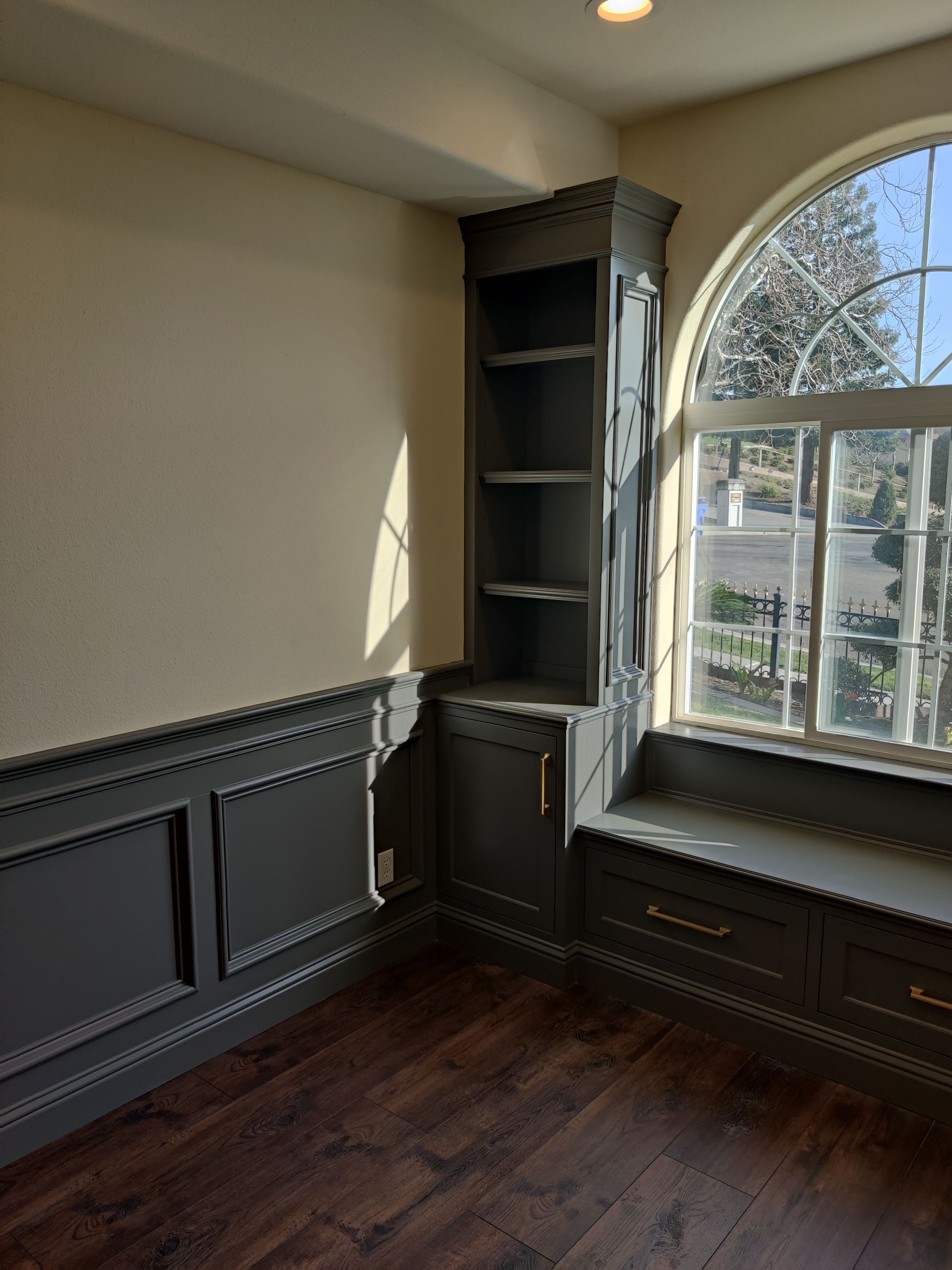 Empty room with dark wood floors, built-in shelves, and a large arched window letting in sunlight.