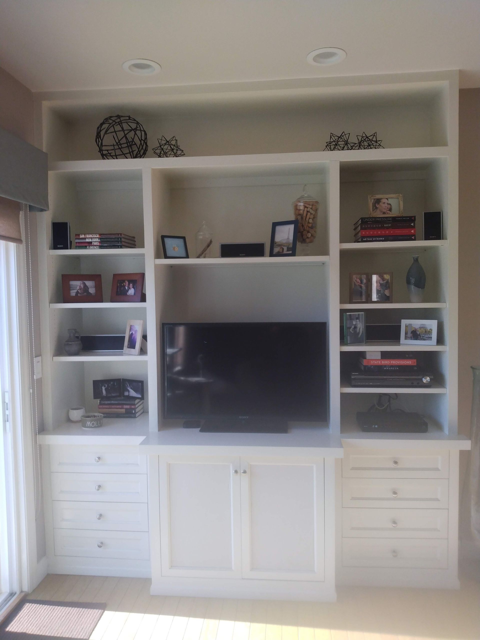 White built-in entertainment center with TV, shelves, cabinets, and decor in a bright living room.