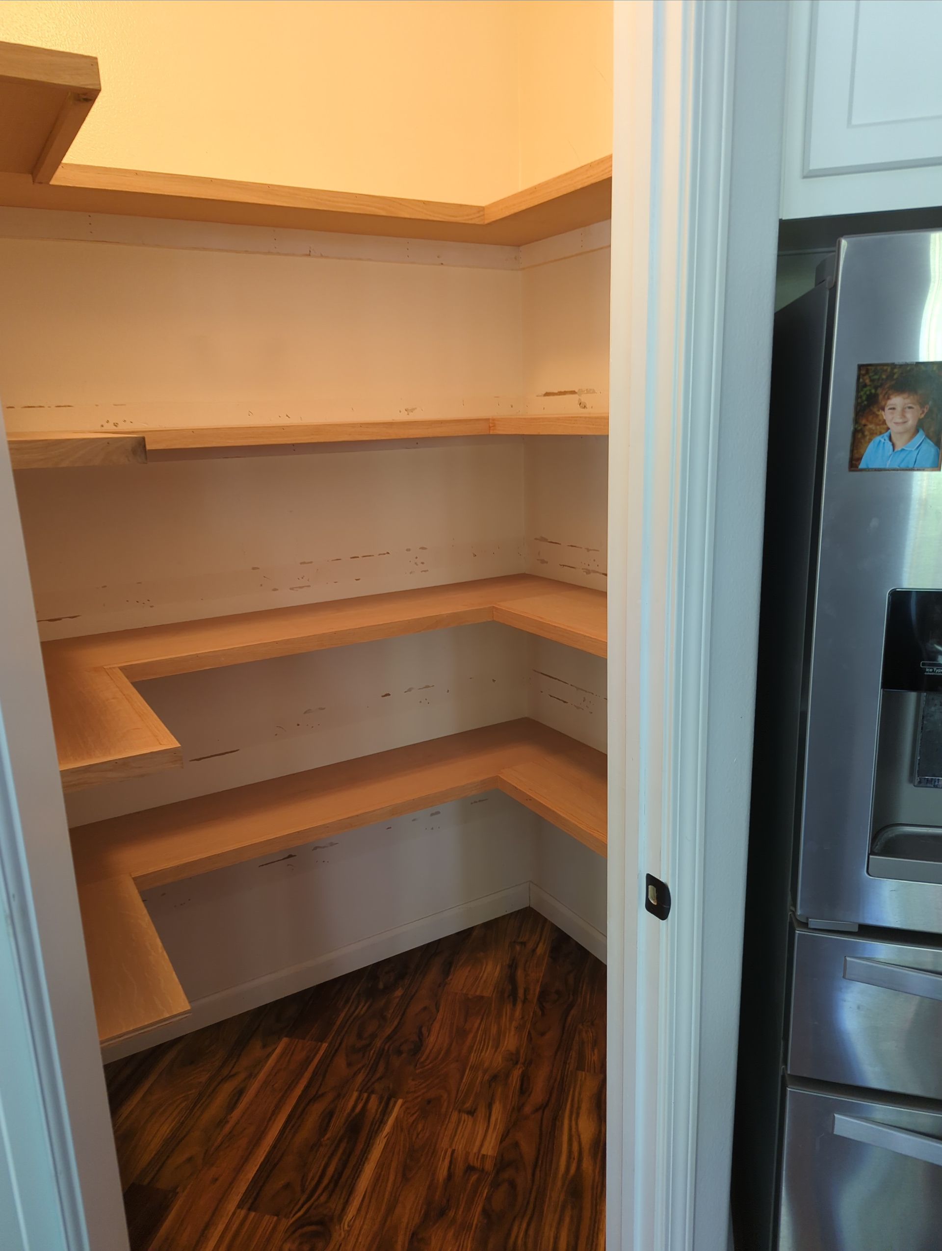Empty pantry with wooden corner shelves, light walls, and dark wood flooring, viewed through a doorway